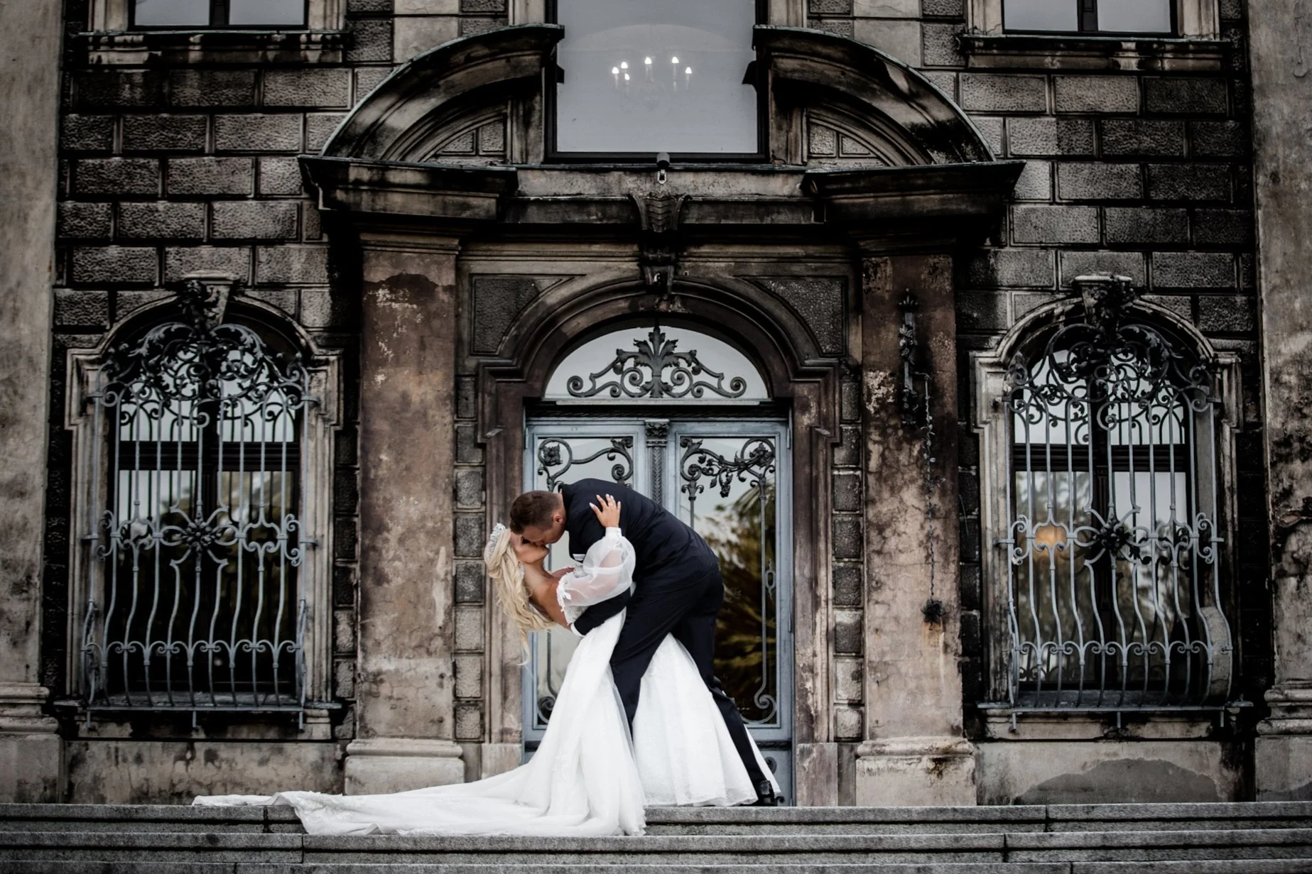 A couple in wedding attire sharing a kiss and dance on the steps in front of an ornate, old building with ironwork windows.