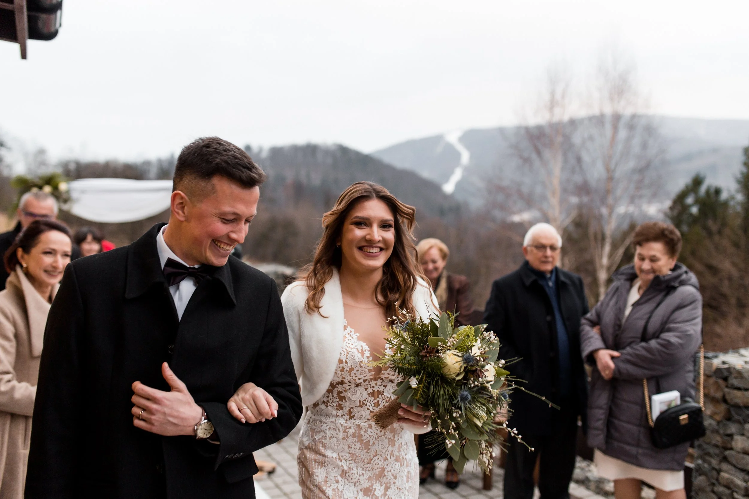 A bride holding a bouquet walks arm-in-arm with her groom at an outdoor wedding ceremony, surrounded by guests with mountains in the background.