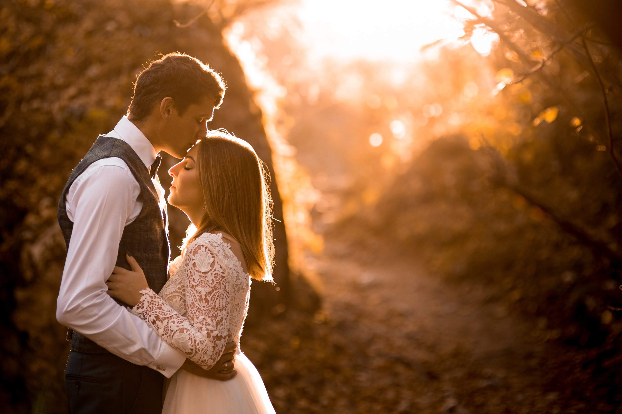 A romantic scene of a couple in formal attire embracing in a wooded area at sunset.