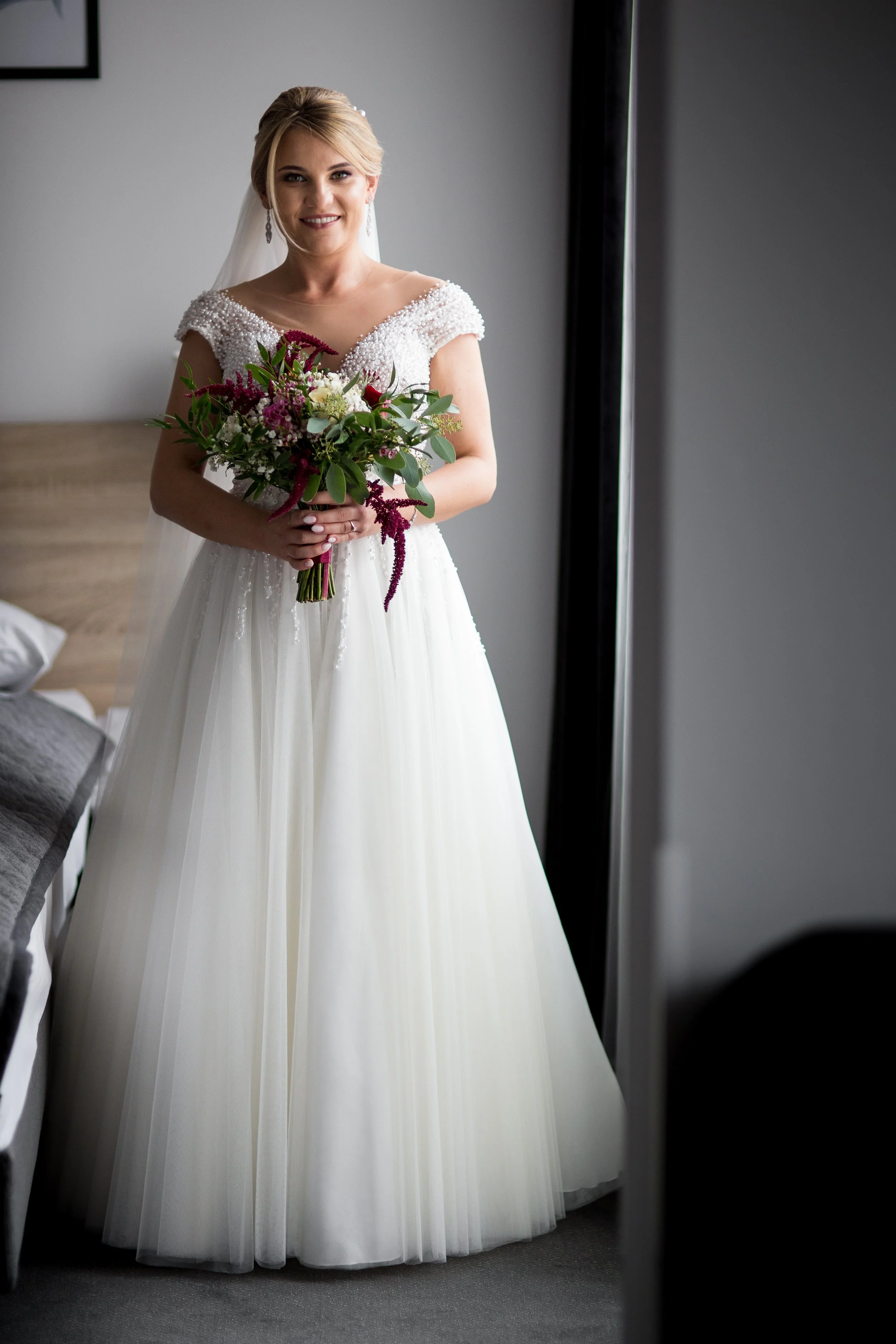 Bride in a white wedding dress holding a bouquet of purple, white, and green flowers, standing indoors near a window.