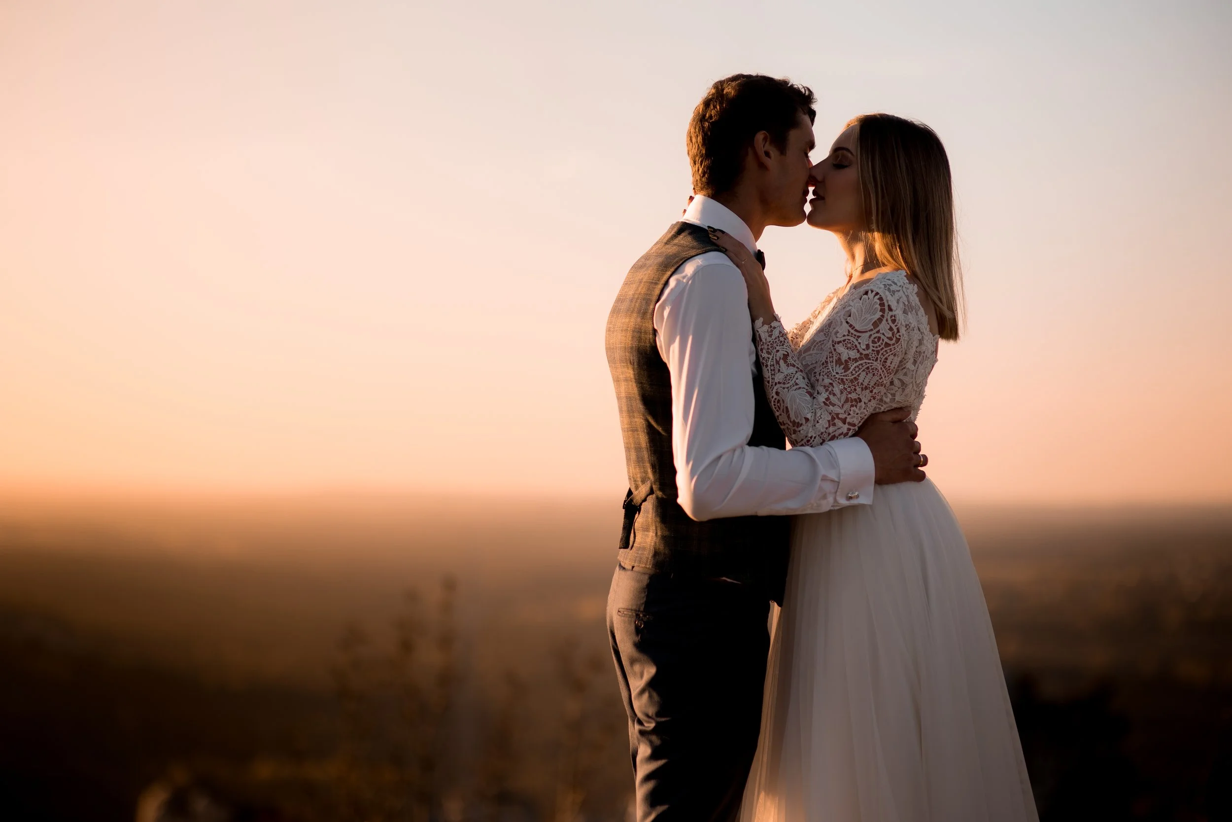 A bride and groom kissing during sunset in an open field, with the sunset sky in the background.