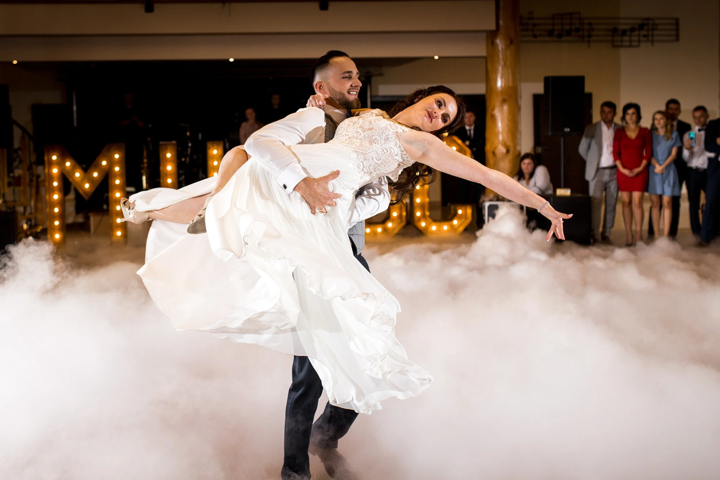 A groom holding a bride in a dance pose on a fog-filled dance floor with wedding guests watching and decorative lights in the background.