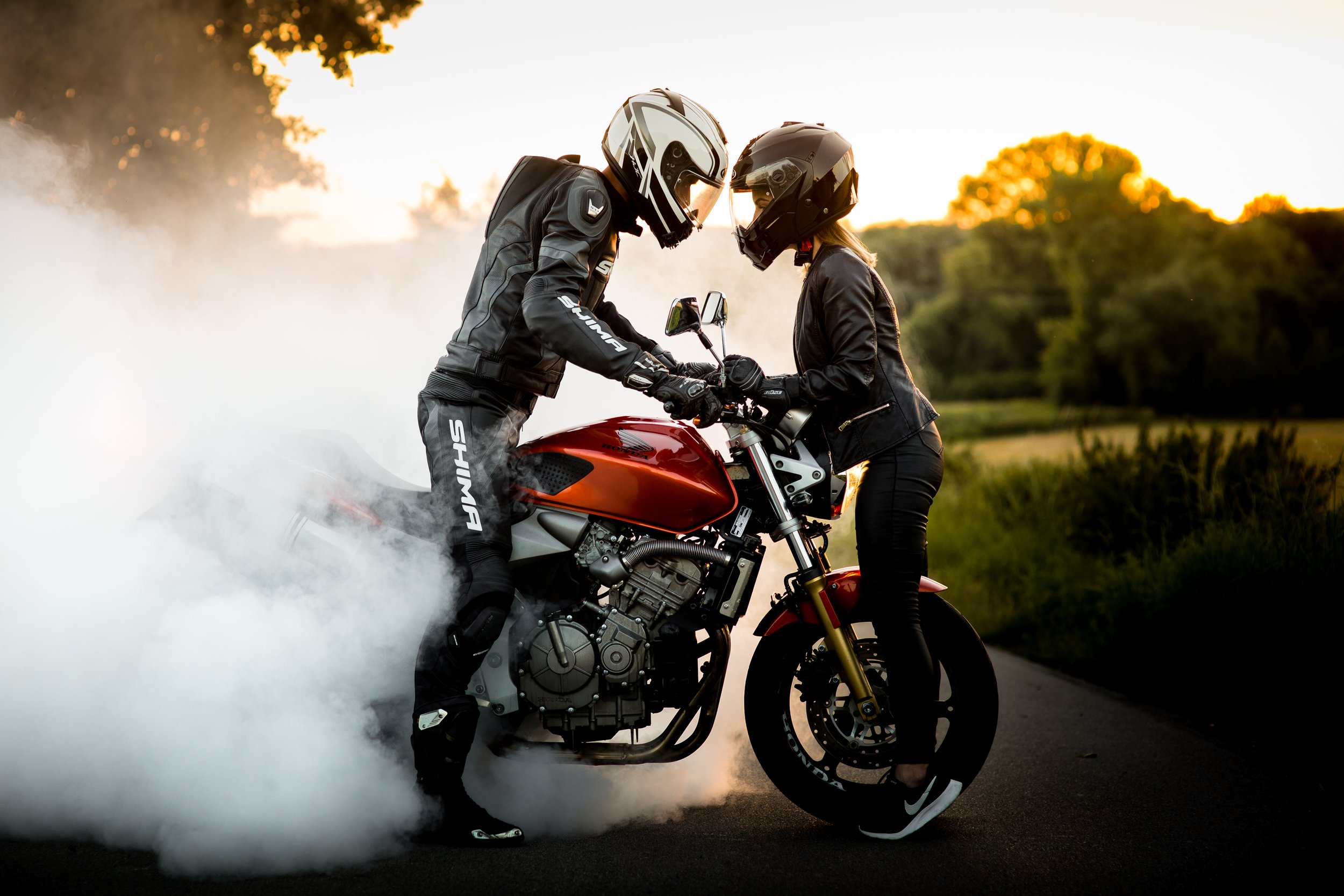 Para zakochanych w strojach motocyklowych, w kaskach i kurtkach, stoją obok czerwonego motocykla na otwartej drodze o zachodzie słońca. Z opon unosi się dym.