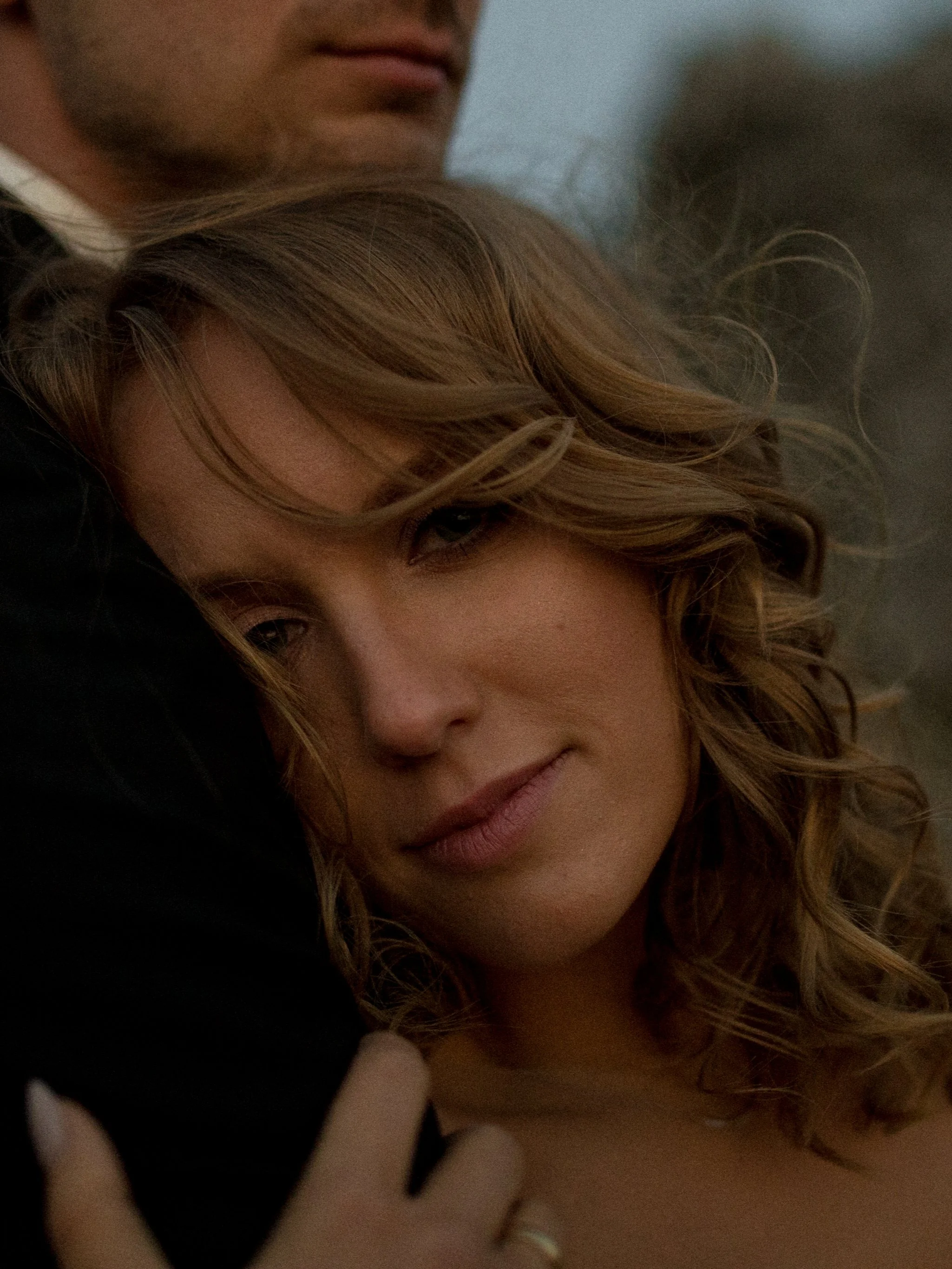 Close-up of a woman with curly blonde hair being embraced by a man, focusing on their faces and upper bodies.