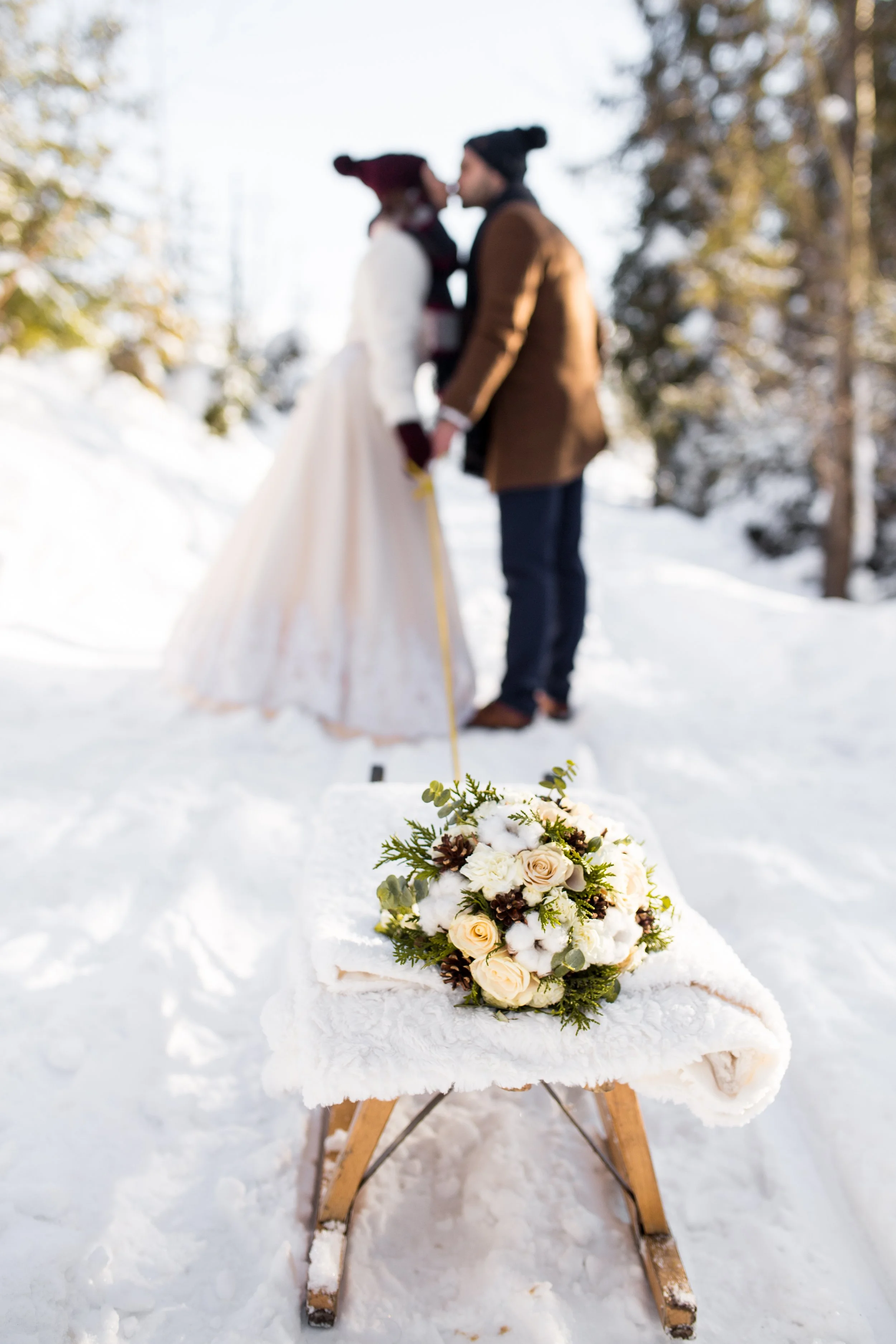 Rozmazana para w strojach ślubnych całująca się na świeżym powietrzu zimą, na pierwszym planie bukiet białych róż i zieleni na małych drewnianych saniach na śniegu.