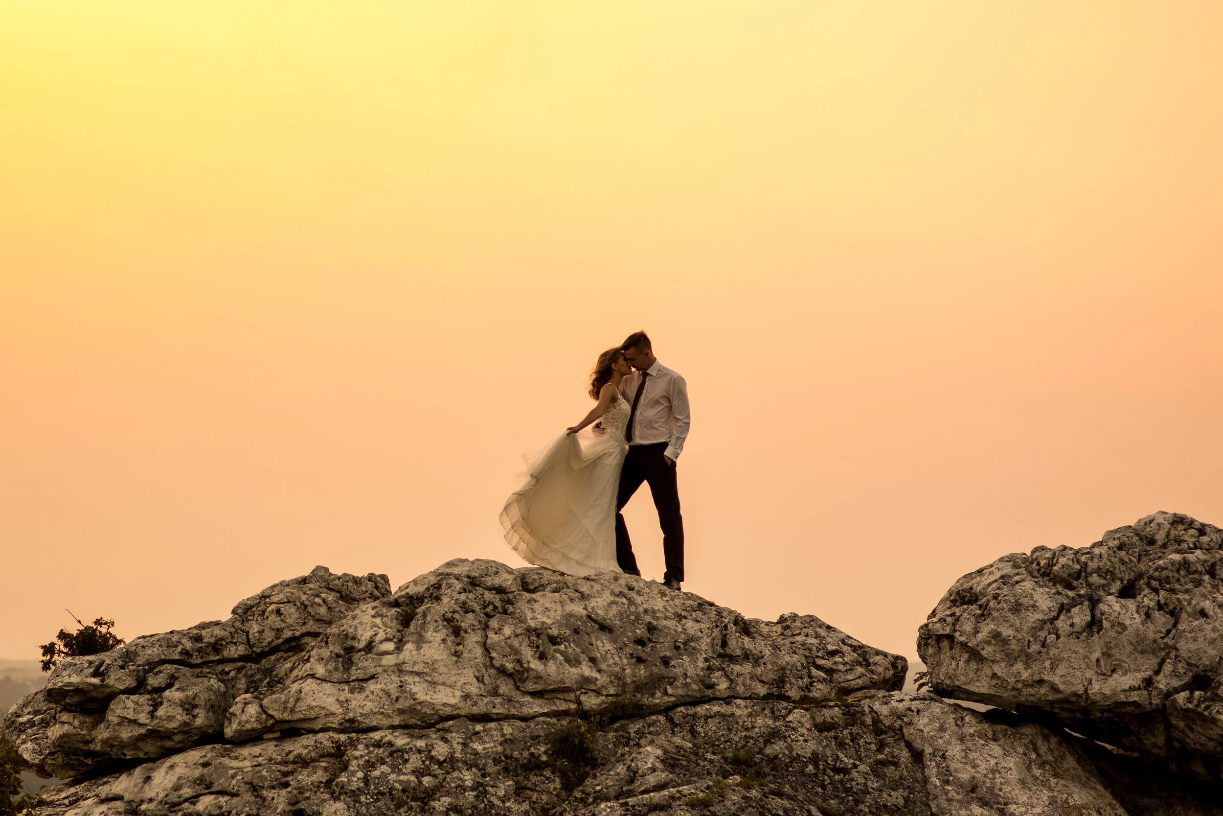 A couple dressed in wedding attire standing on a rocky mound during sunset, sharing an intimate moment.