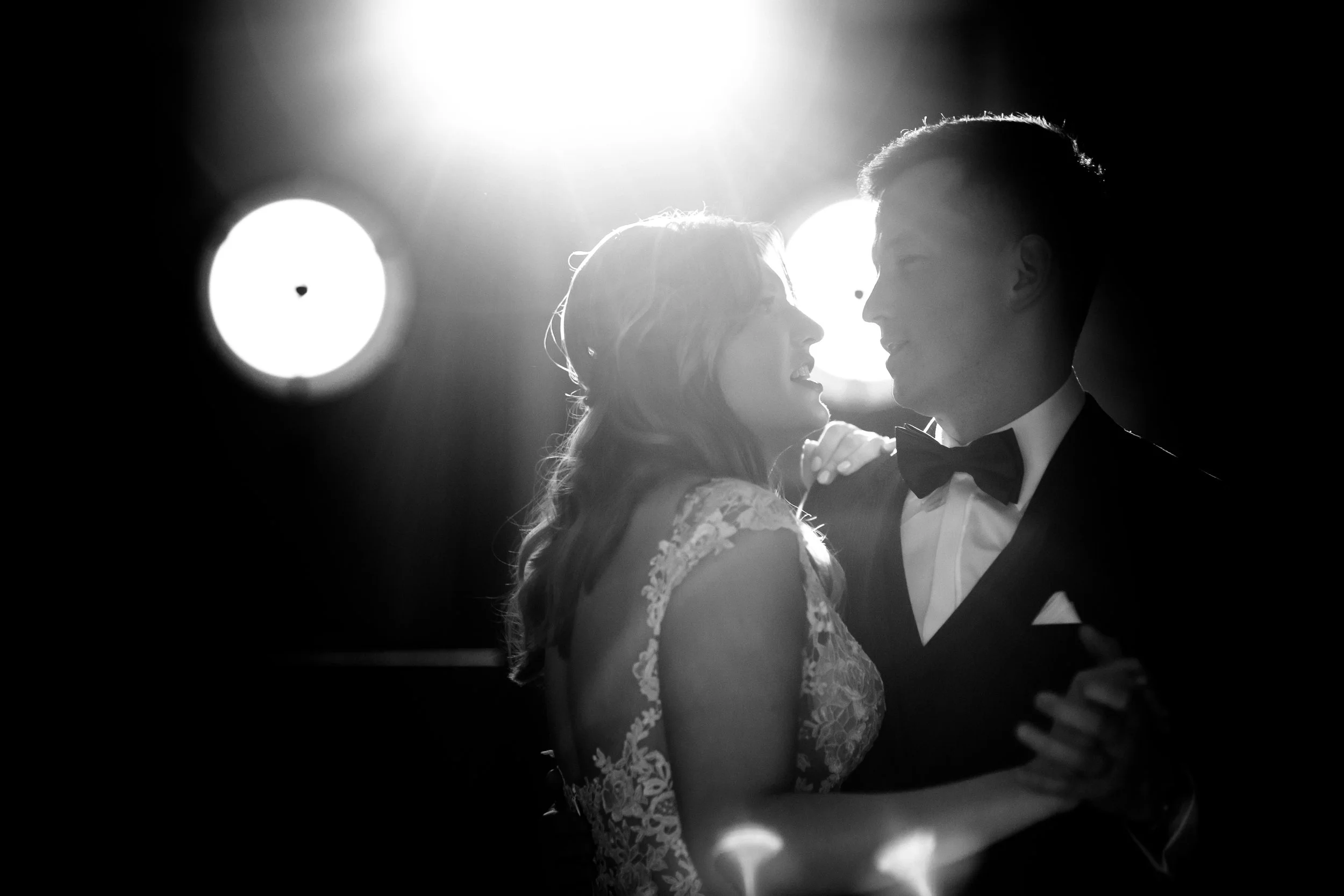 A black and white photo of a couple dancing closely, backlit with bright lights, the woman in a lace dress and the man in a tuxedo with a bow tie.