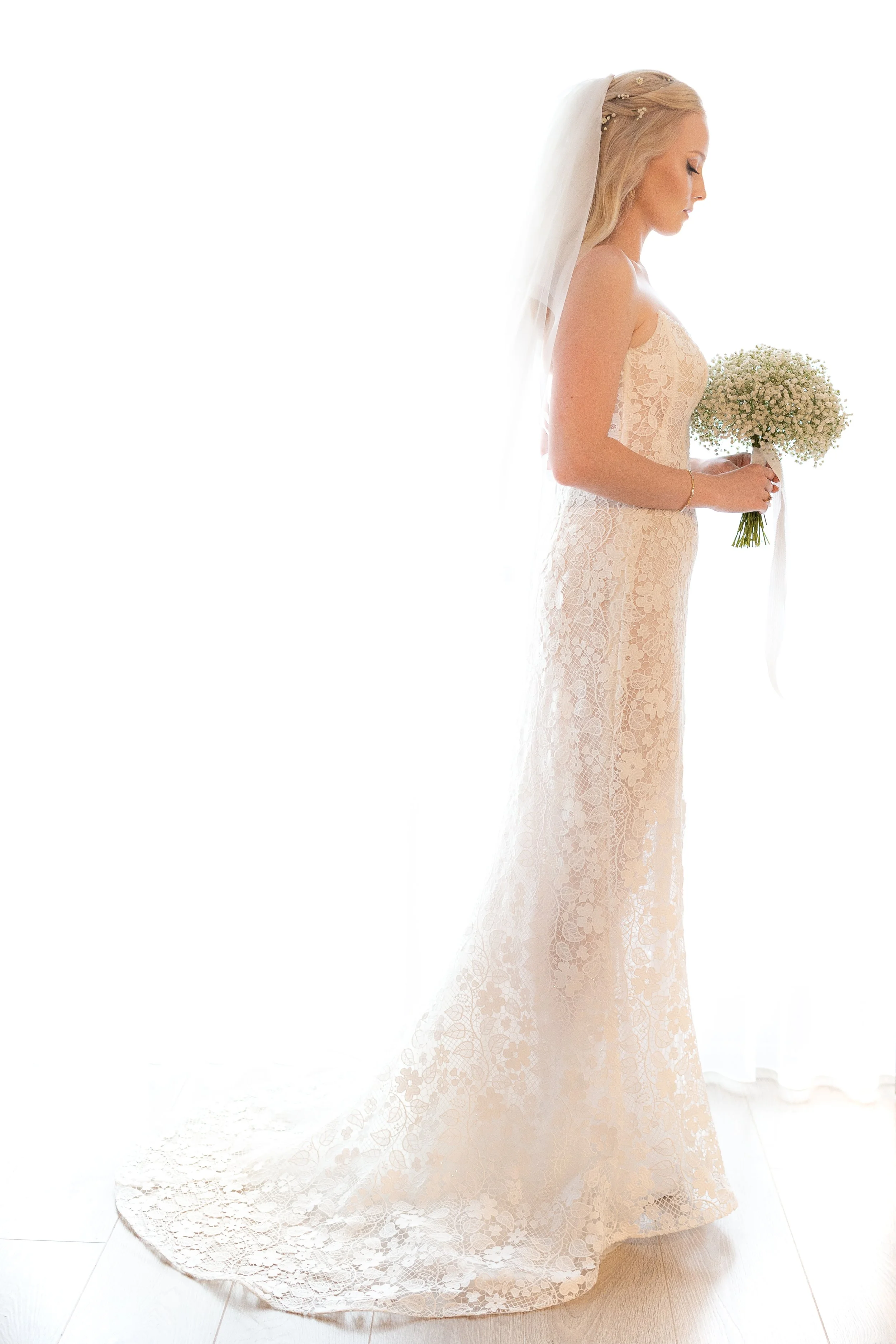 A bride in a lace cream-colored wedding gown holding a bouquet of white flowers, standing in profile with her eyes closed, wearing a veil and jewelry, near a white curtain.