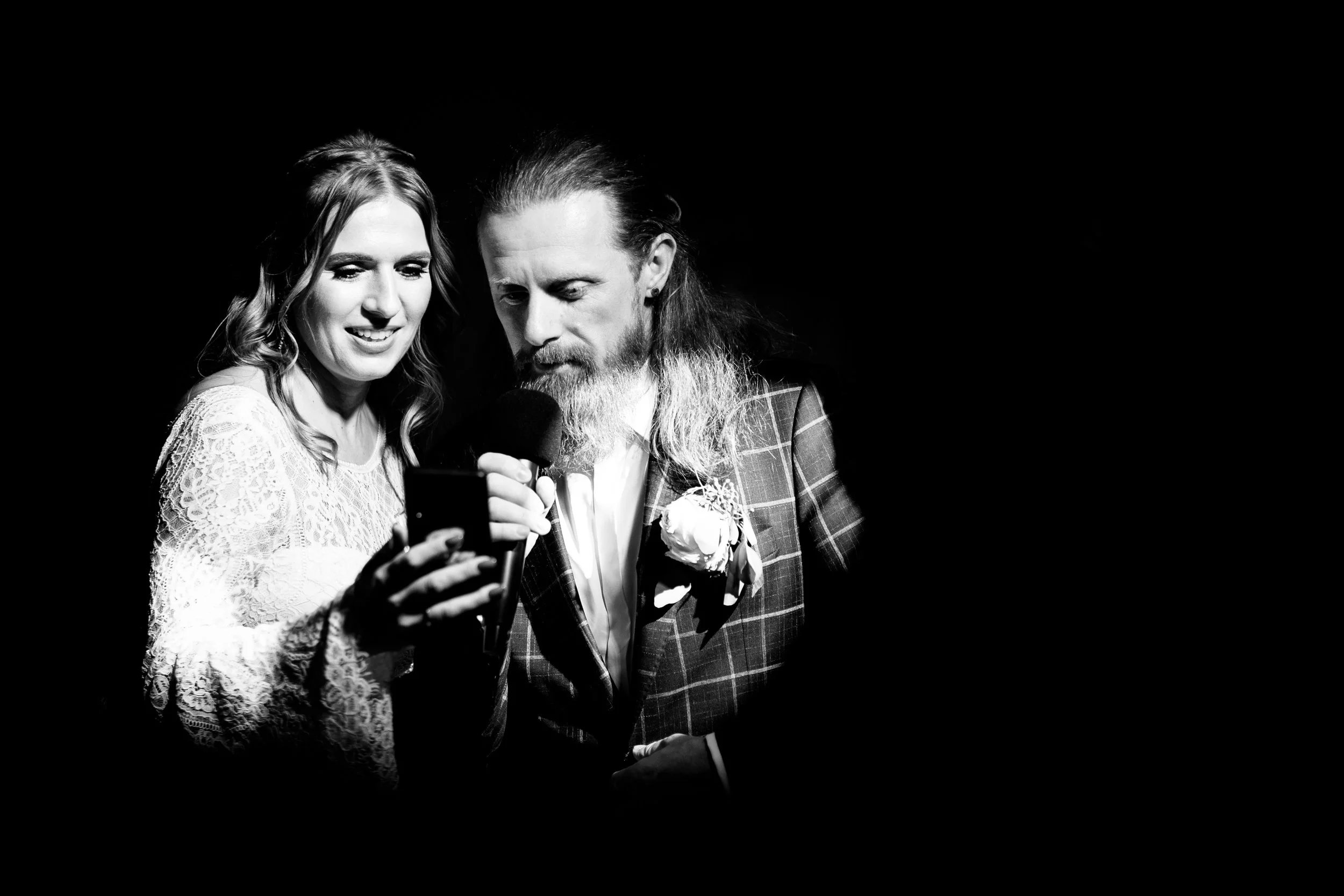 A bride and groom looking at a phone while speaking into a microphone during their wedding celebration, with the bride smiling and wearing a lace dress, and the groom in a plaid suit with a boutonnière.