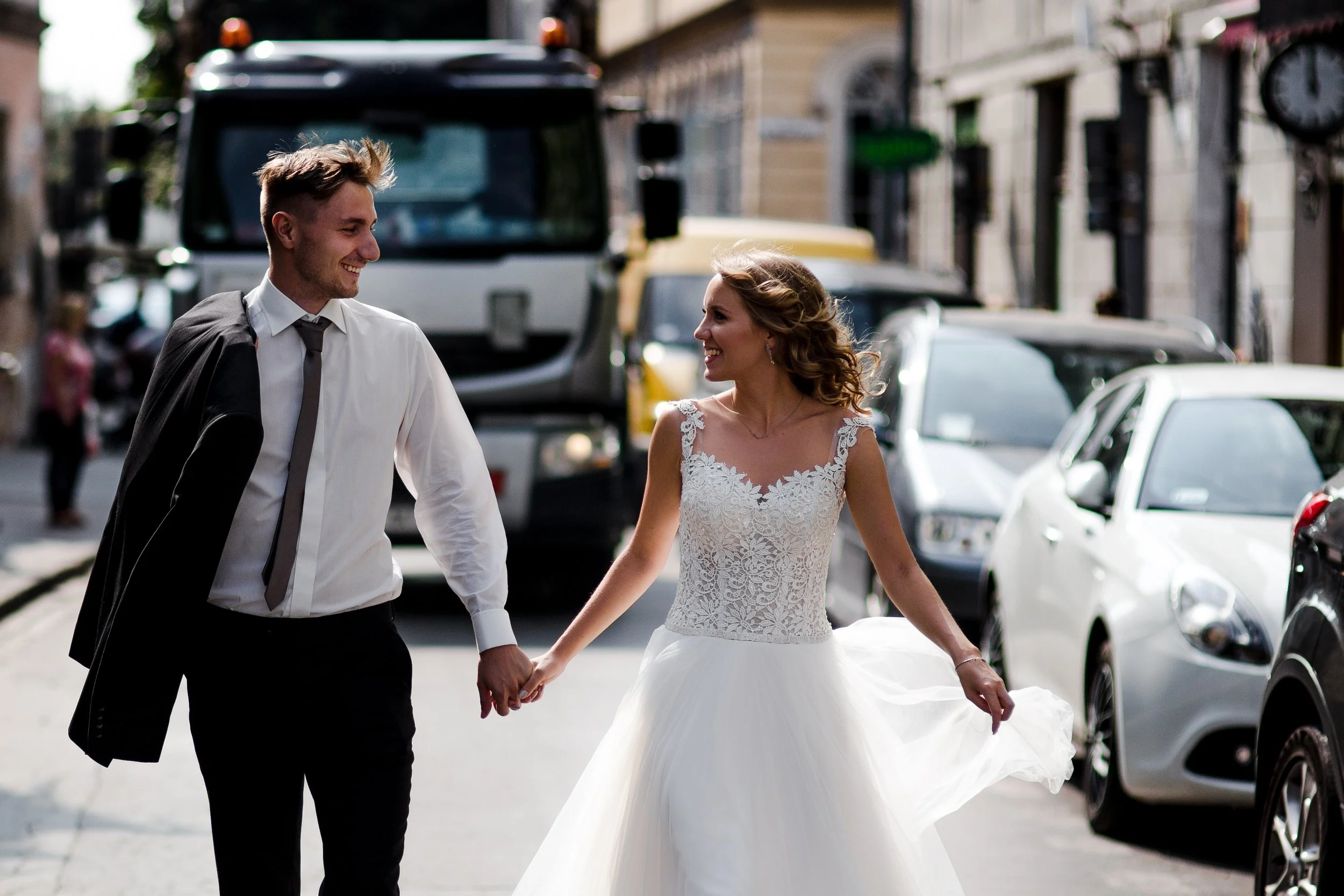 A newlywed couple holding hands, walking on a city street, smiling at each other, with cars parked along the curb and buildings in the background.