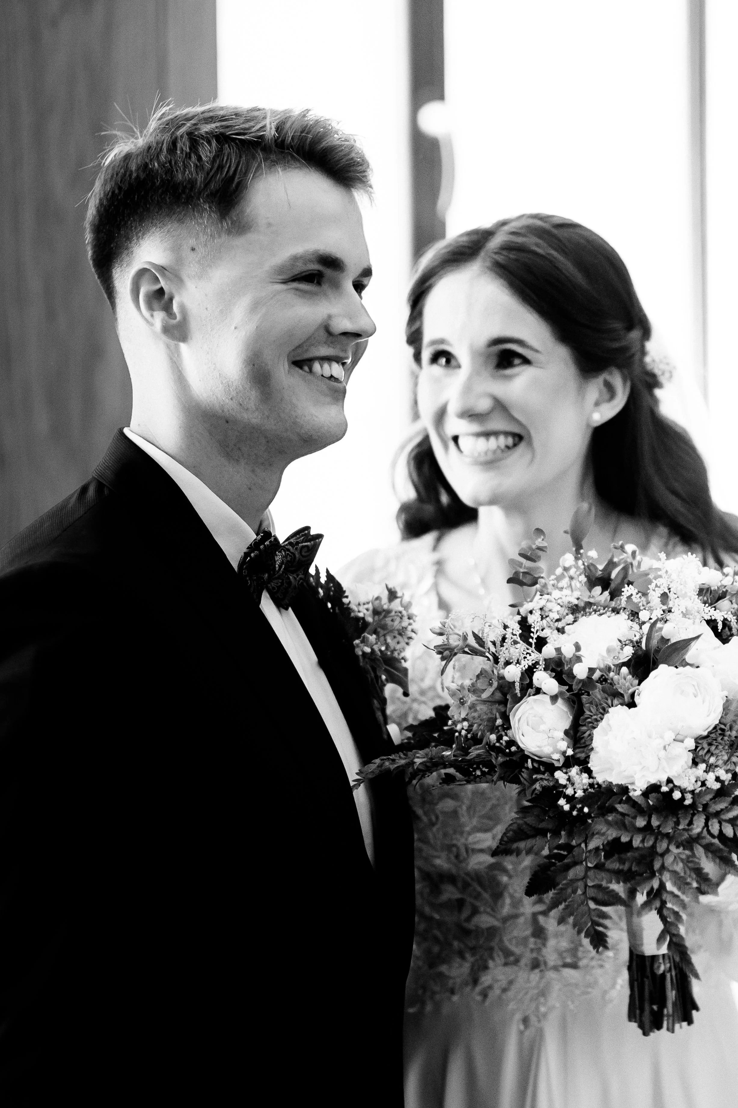 Czarno-białe zdjęcie uśmiechniętej panny młodej trzymającej bukiet i pana młodego w smokingu na ślubie.