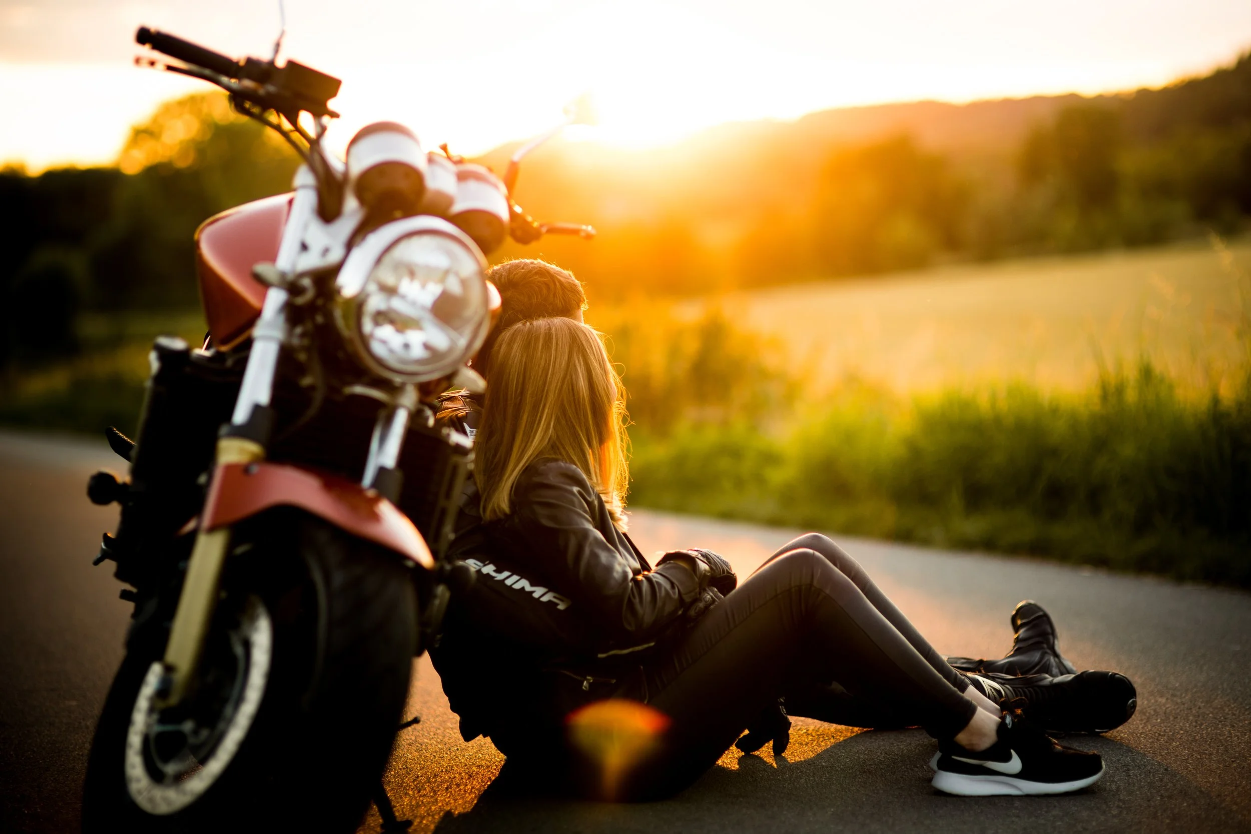 Dwie osoby siedzą na drodze obok motocykla podczas zachodu słońca, w tle widać wiejski krajobraz.