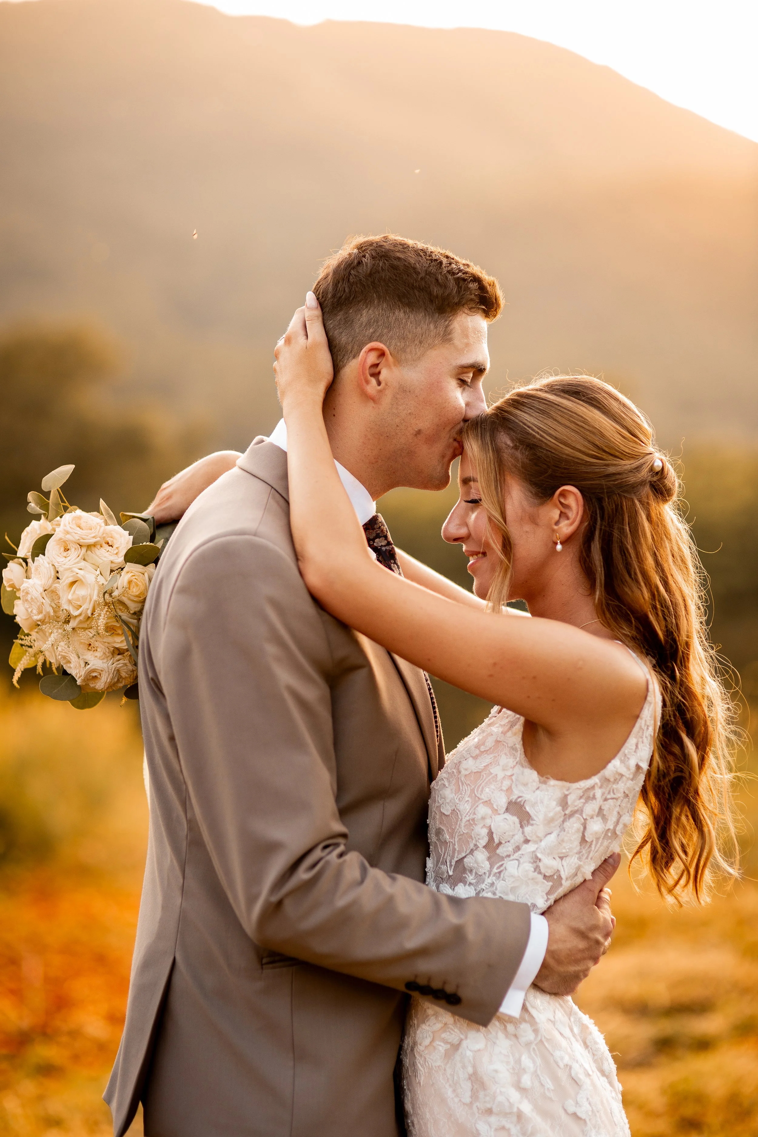 A newlywed couple sharing a kiss outdoors during sunset, with the groom in a beige suit and the bride in a lace wedding dress, holding a bouquet of white roses.