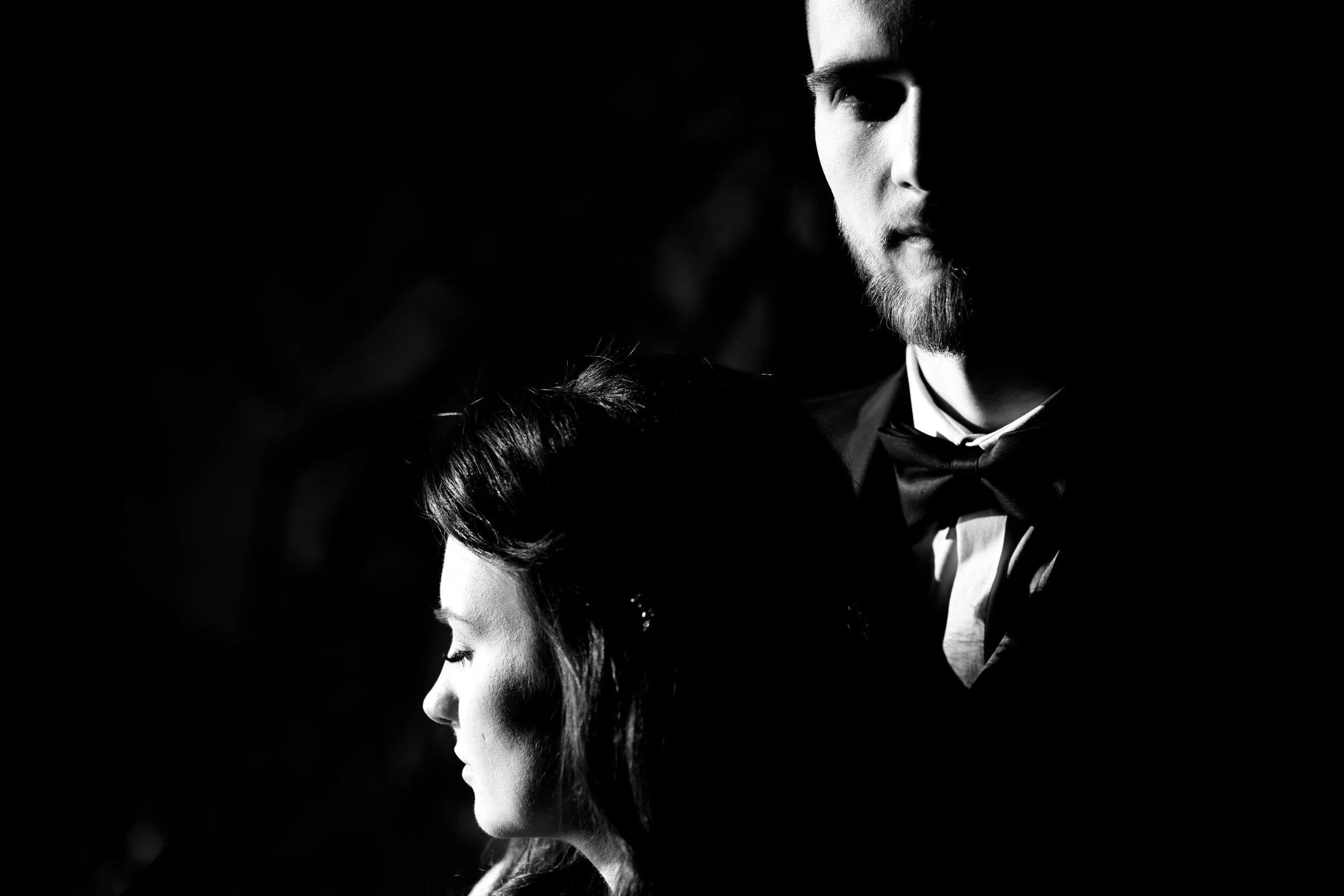 A black and white photo of a man and woman, with the woman's face in profile and the man's face partially visible. The woman has dark hair styled in vintage waves, and the man has a beard, short hair, and is wearing a tuxedo with a bow tie. The light