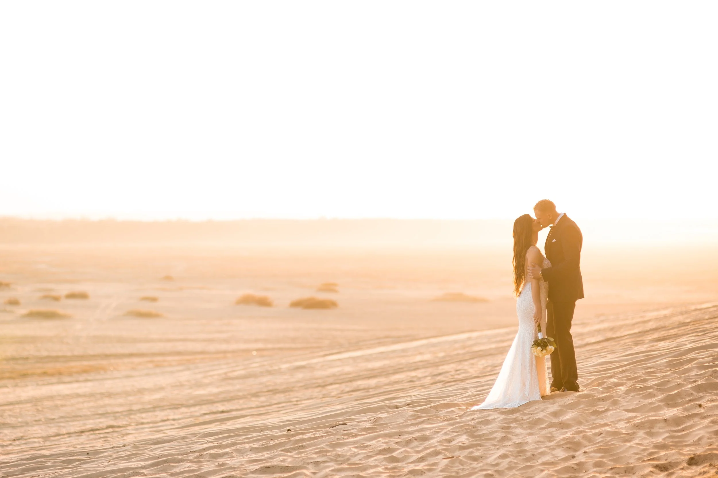 A bride and groom embrace on a sandy beach at sunset, with the bride holding a bouquet of flowers.