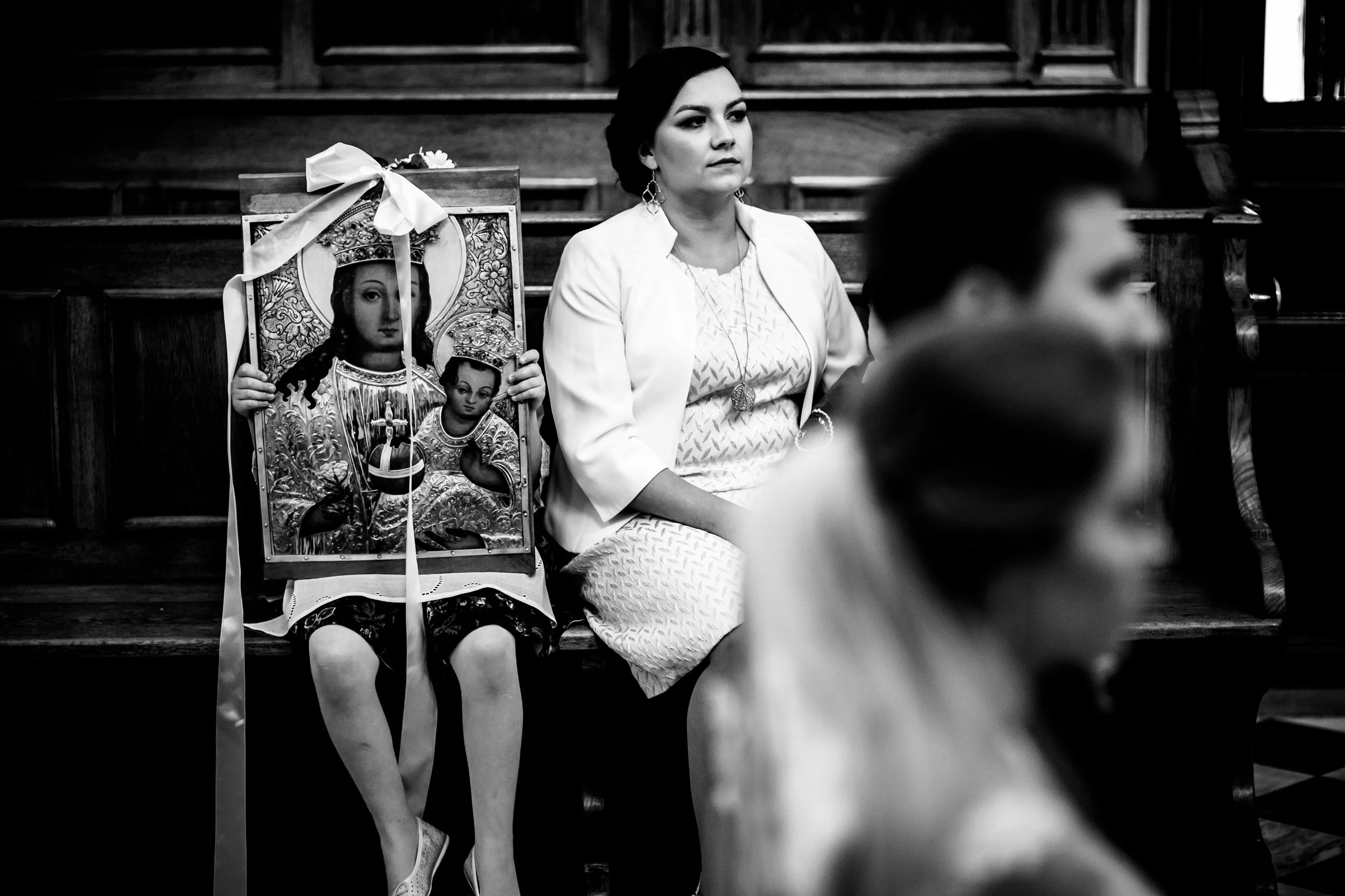 A woman sitting in a church pew with a child behind her holding a religious icon of the Virgin Mary and baby Jesus. The woman has dark hair, wearing a white dress and white blazer, and appears to be attending a religious event or service. The image i