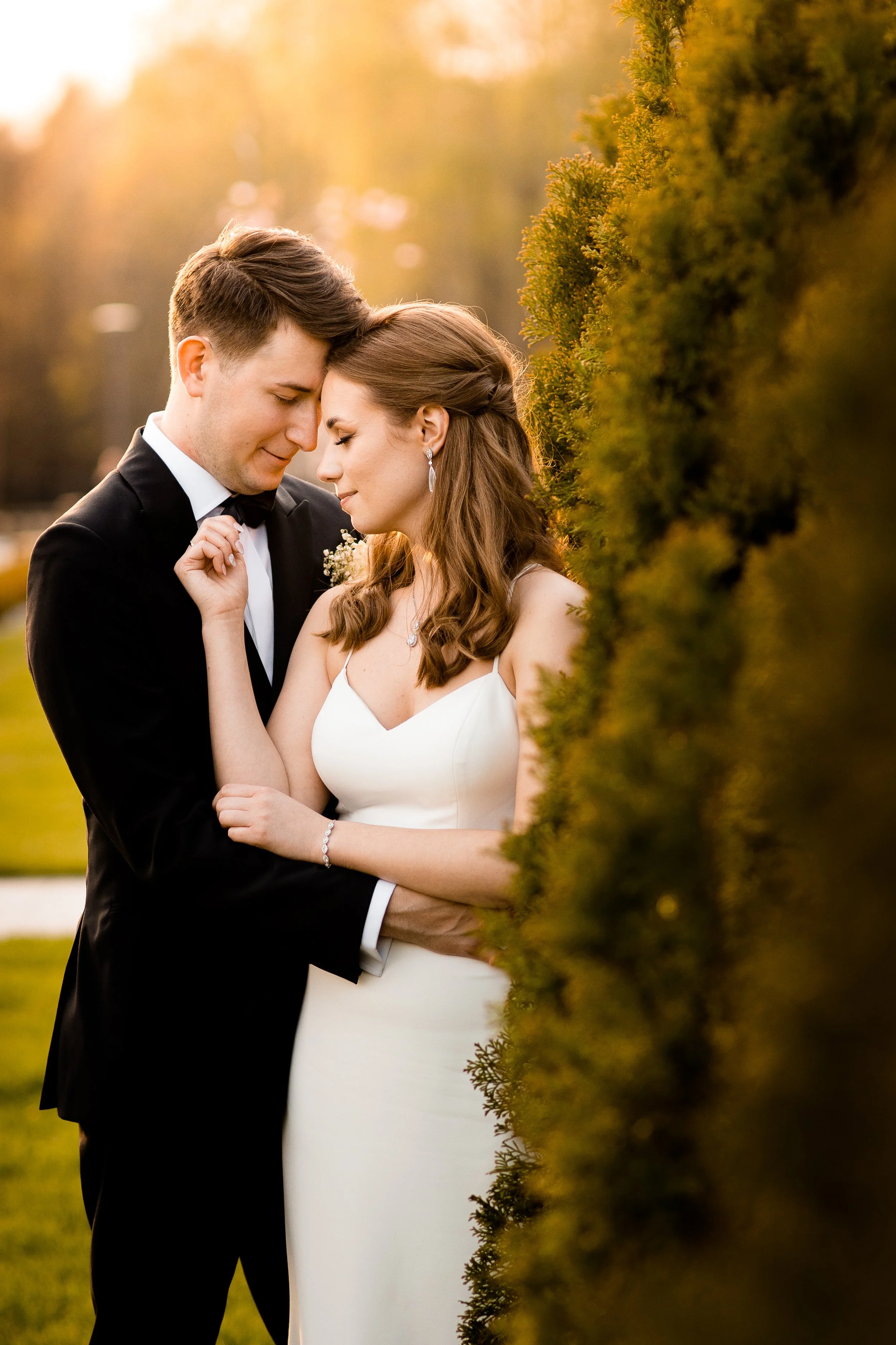 A bride and groom standing close with foreheads touching, smiling, outdoors during sunset.