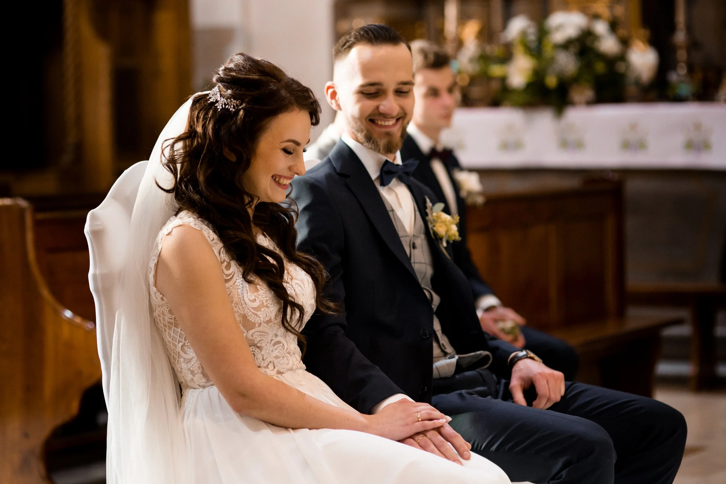 A bride and groom sitting in a church during their wedding ceremony, holding hands and smiling, with other guests visible in the background.