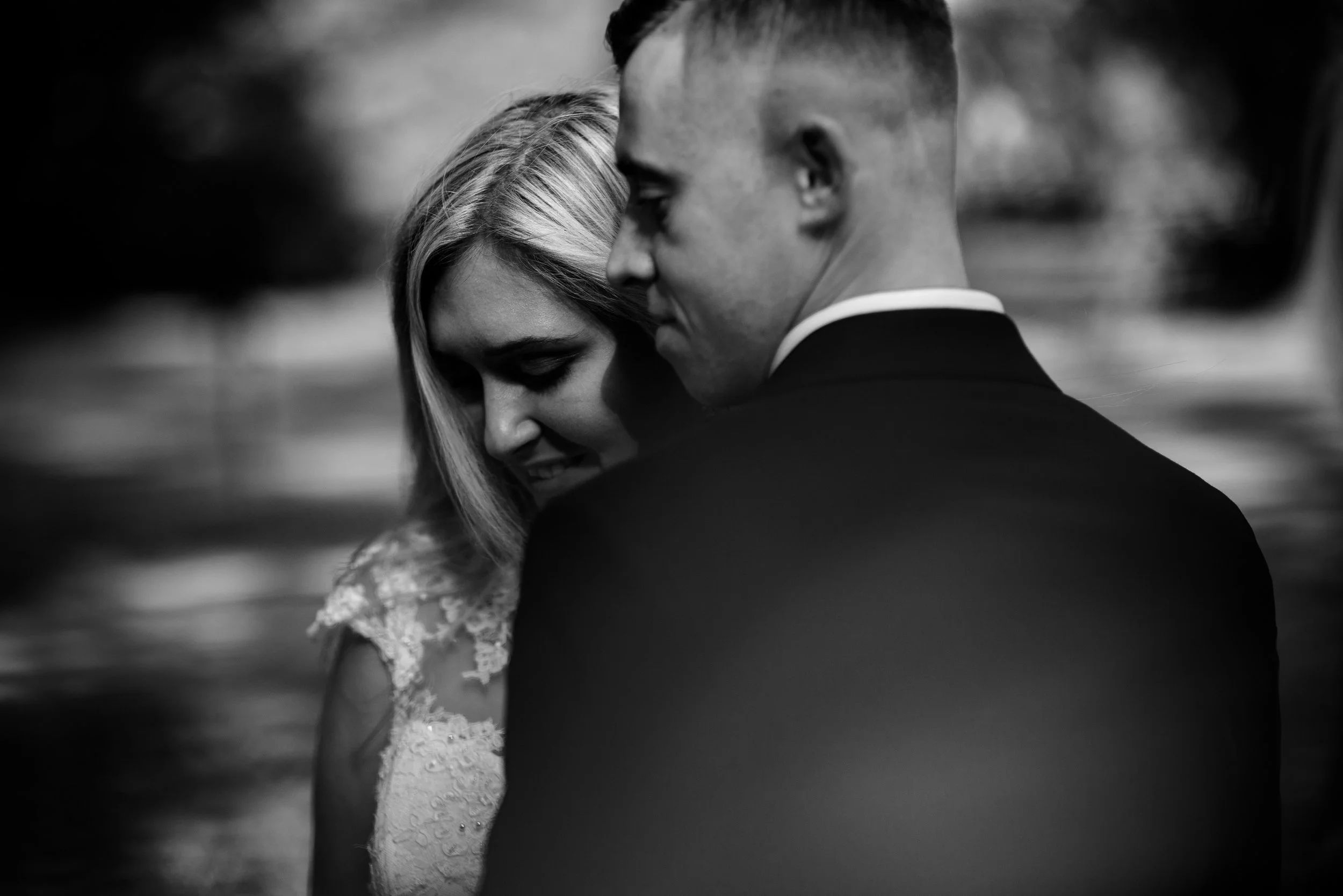 A black-and-white photo of a couple embracing, with the woman smiling with her eyes closed and the man facing away from the camera, wearing a suit.