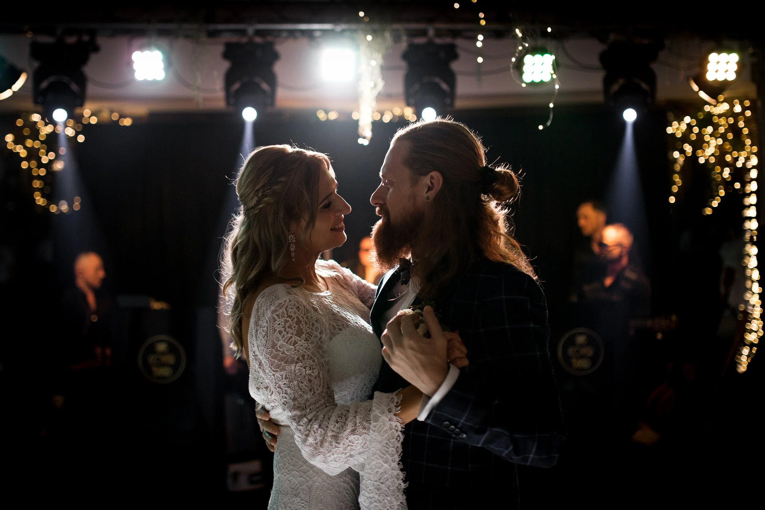 A bride and groom dancing closely at their wedding reception, illuminated by soft lighting and surrounded by string lights and guests in the background.