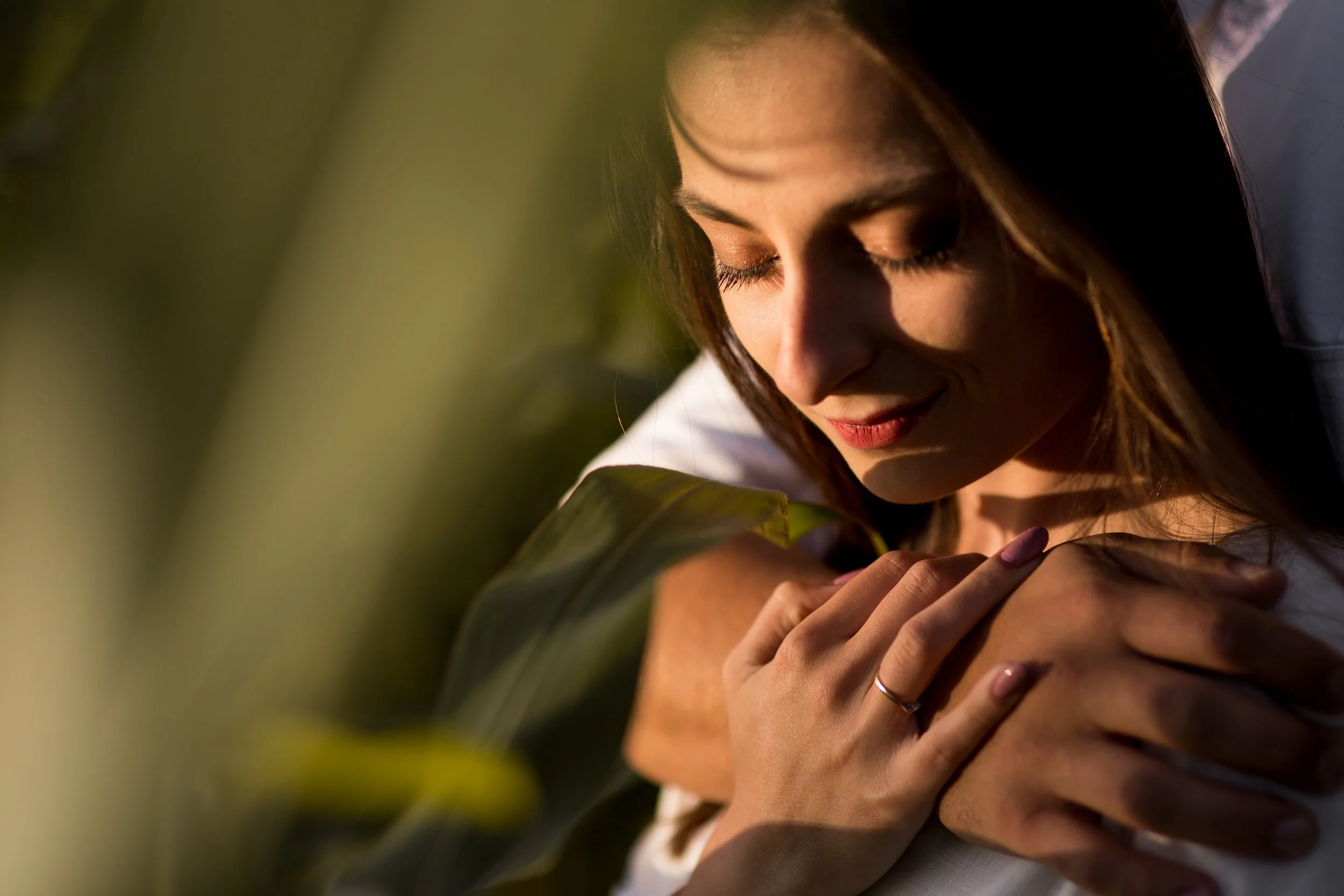 A woman with long brown hair and closed eyes gently holds a large green leaf close to her face outdoors in warm, natural light.