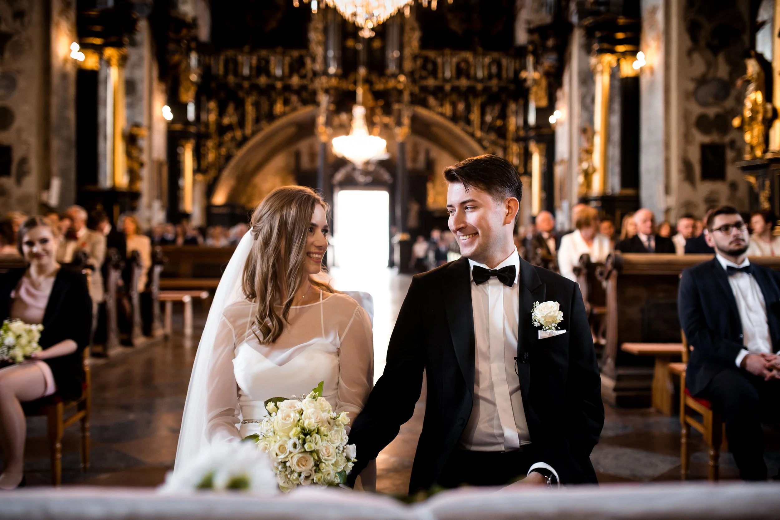 A bride and groom at their wedding ceremony inside a church, both smiling and looking at each other, with wedding guests seated behind them.
