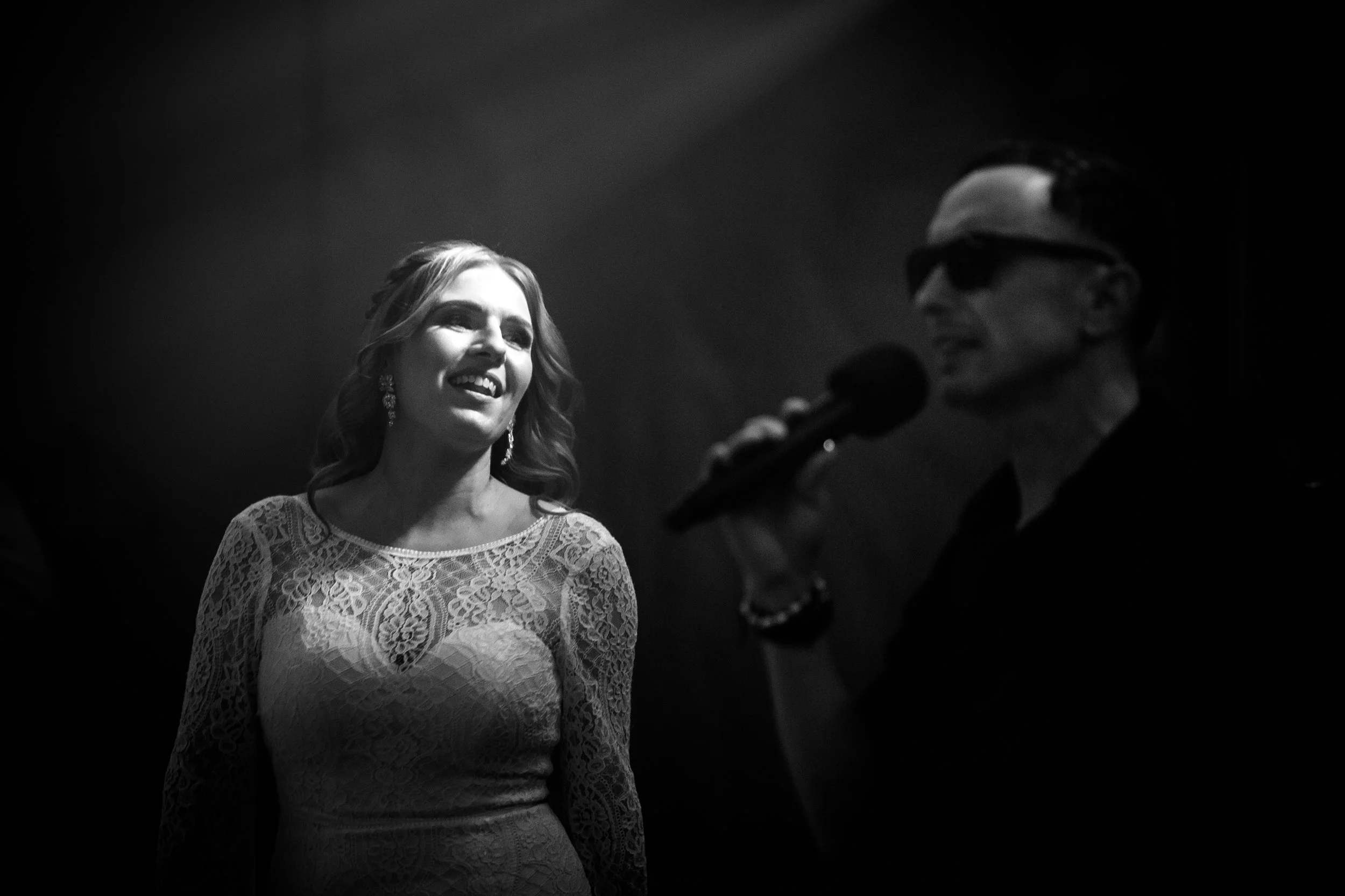 A woman in a lace dress smiling while looking at a man holding a microphone, who is wearing sunglasses and singing.