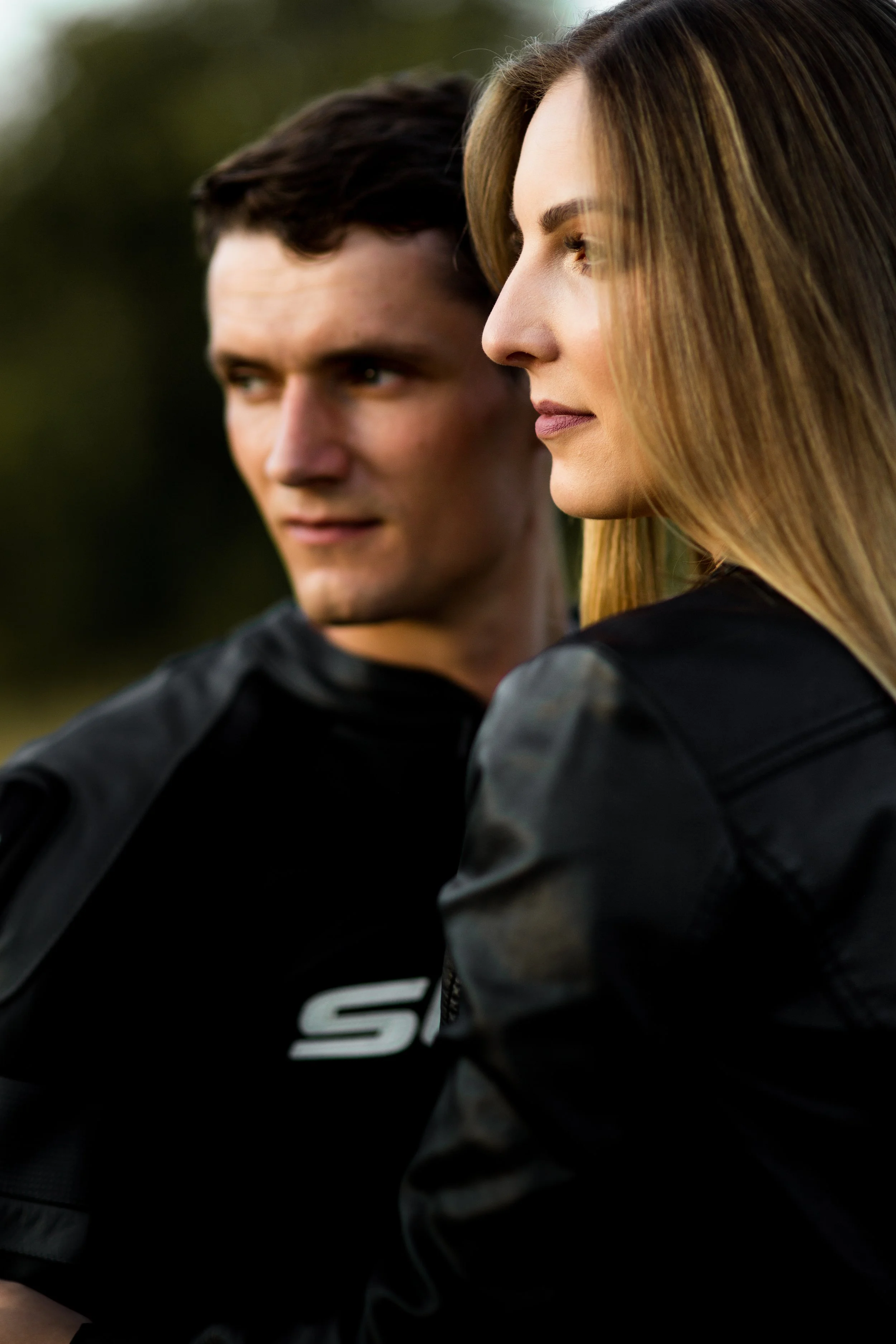 A man and woman in black leather jackets outdoors, looking pensively into the distance at sunset.