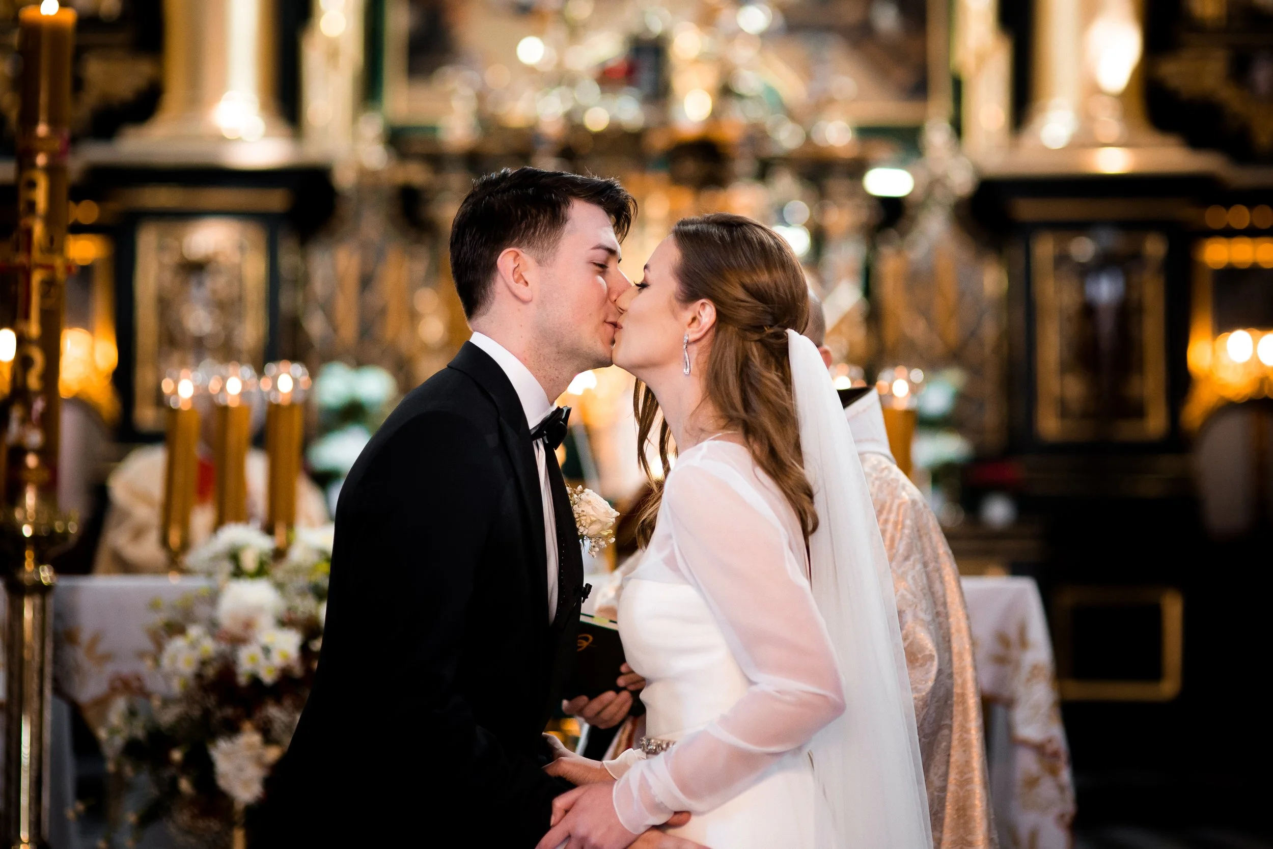 Para młoda całuje się podczas ceremonii ślubnej w kościele, w tle widać świece i kwiaty.