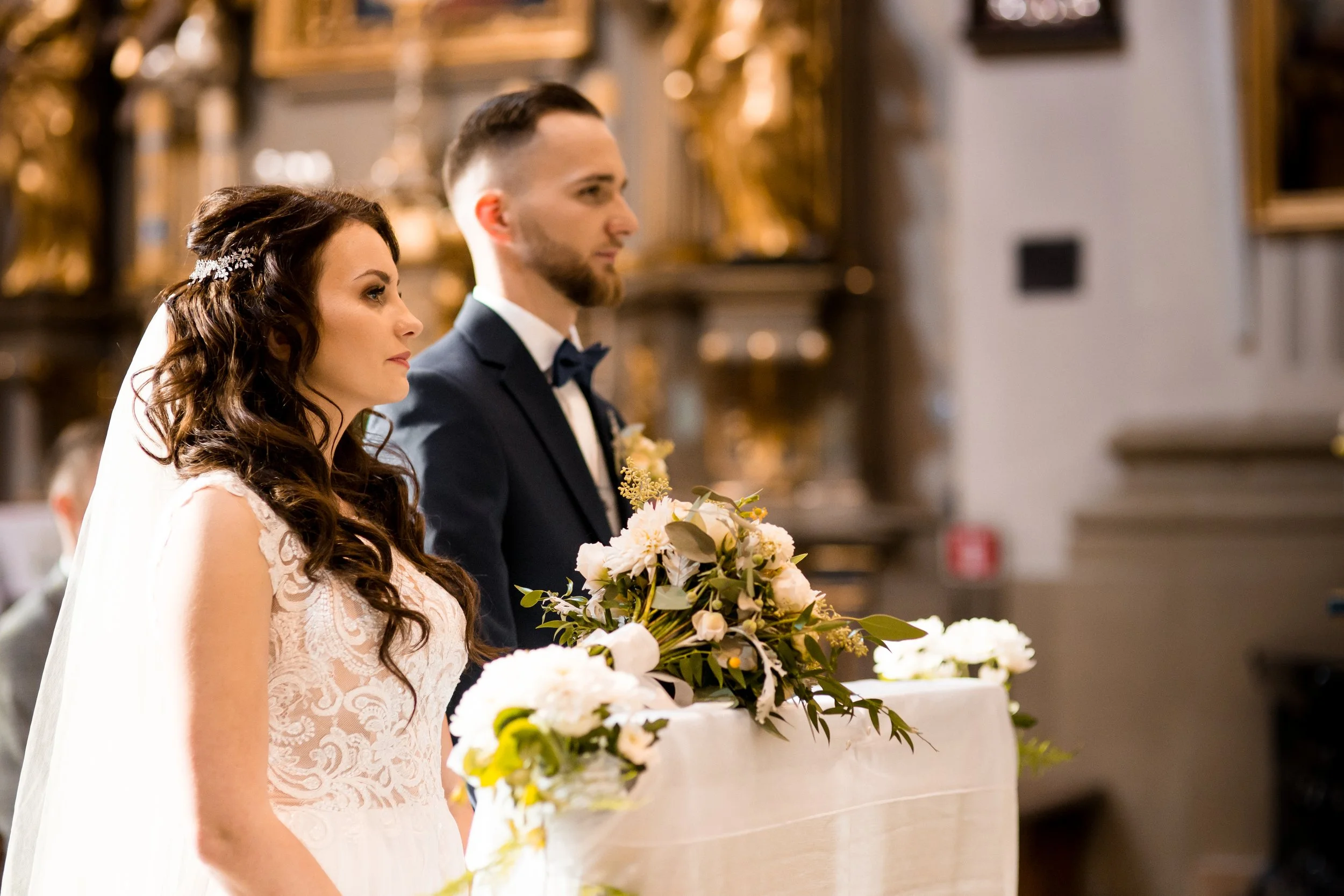 A bride and groom standing side by side during wedding ceremony, with the bride in a lace wedding dress and the groom in a dark suit and bow tie, in a church or chapel decorated with floral arrangements.
