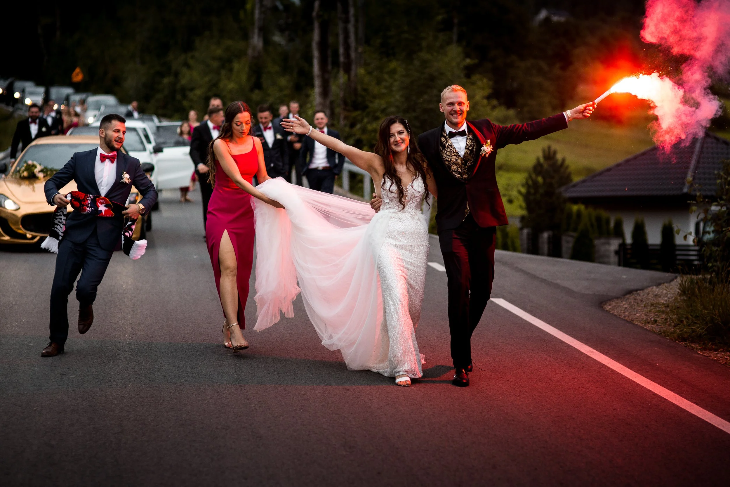 A wedding celebration with a newlywed couple walking on a road, the groom holding a flare, and attendants dressed in formal attire, some holding items like a scarf or a dress train, with parked cars and trees in the background.