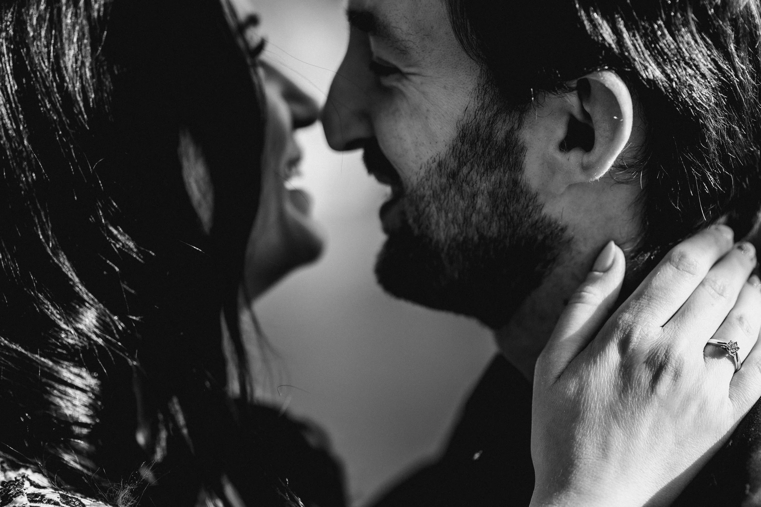 Czarno-białe zdjęcie z bliska przedstawiające parę z twarzami blisko siebie, uśmiechniętą, z dłonią kobiety delikatnie dotykającą twarzy mężczyzny, na której widać pierścionek zaręczynowy.