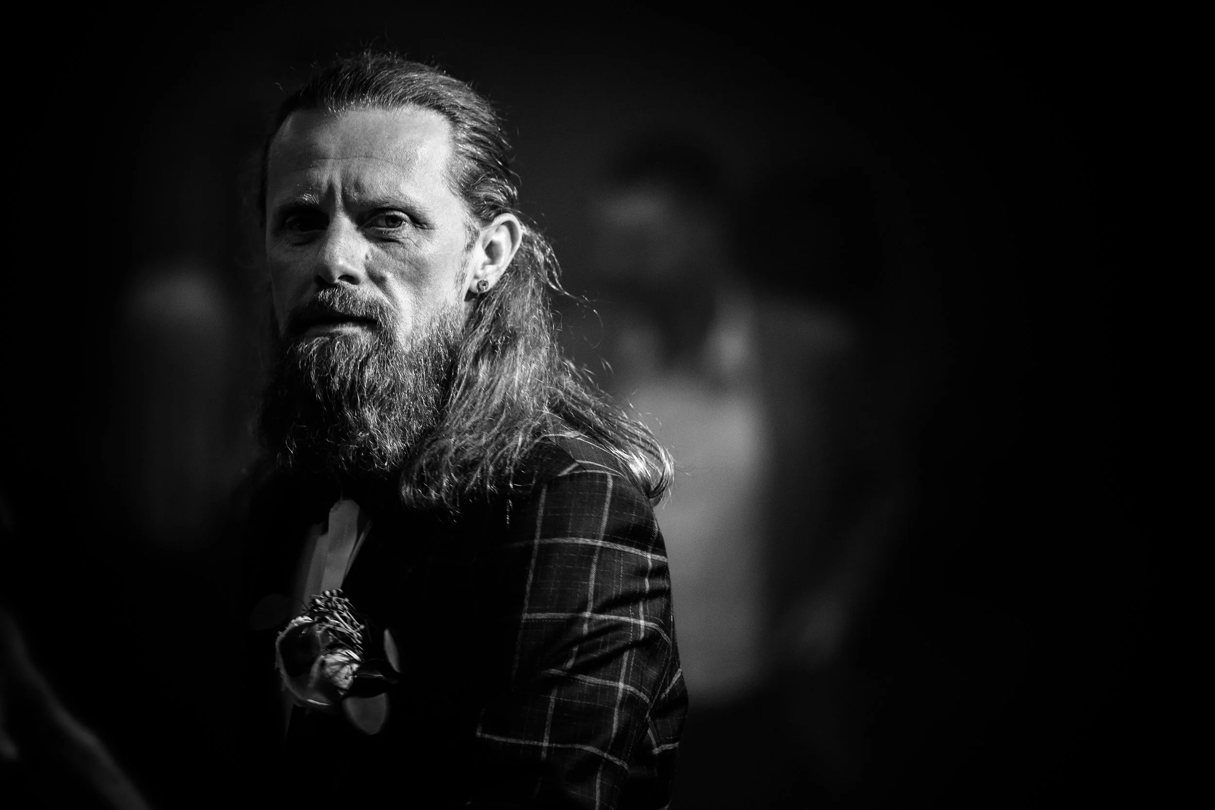 A black and white photo of a man with long hair and a beard, wearing a checkered jacket, looking at the camera with a serious expression, with a blurred background.