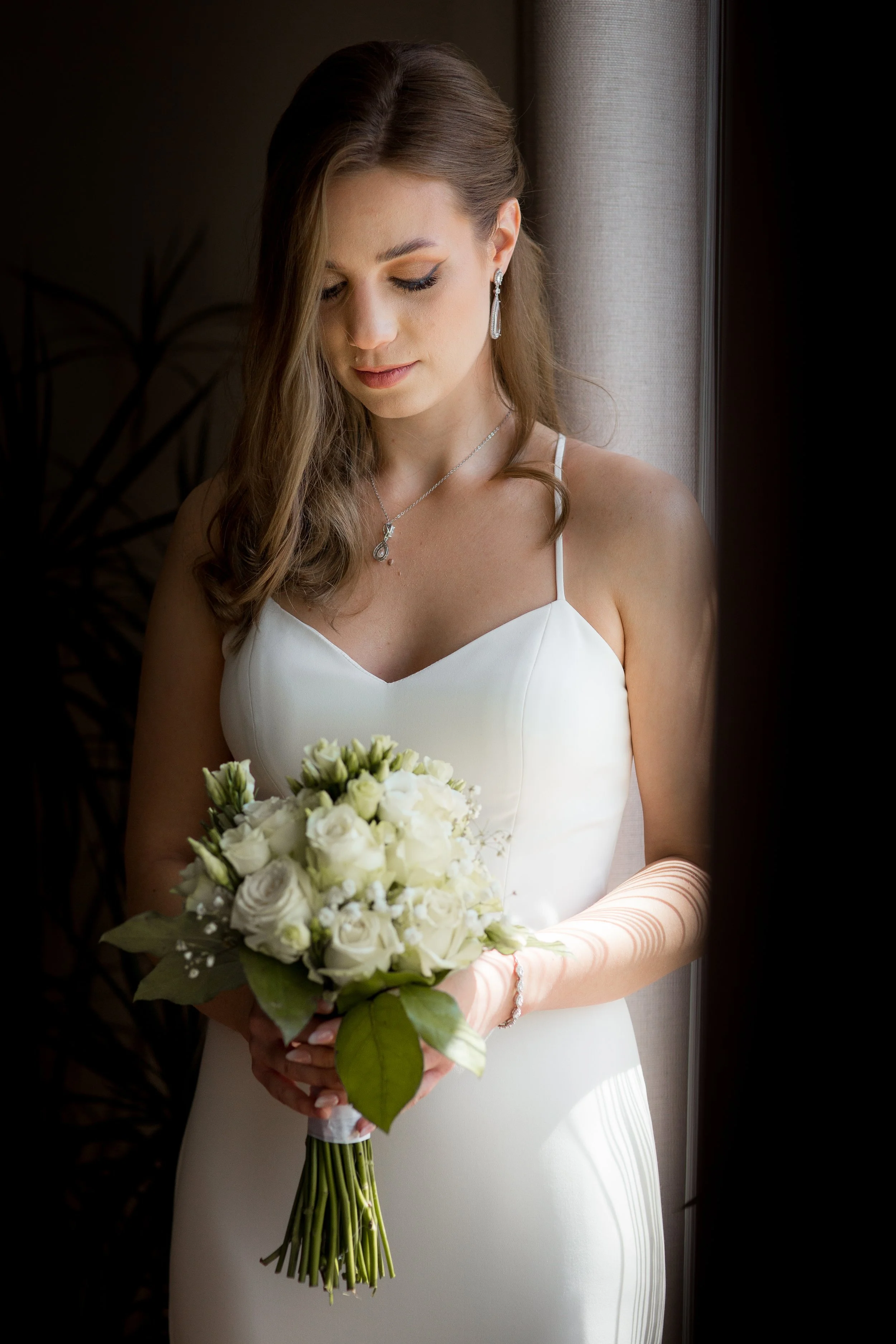 A bride in a white wedding dress holding a bouquet of white roses and greenery, standing near a window with soft natural light.