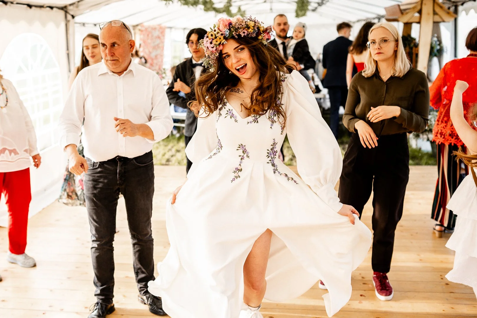A woman wearing a white dress with floral embroidery and a flower crown is dancing and smiling at a celebration or party, surrounded by other people.