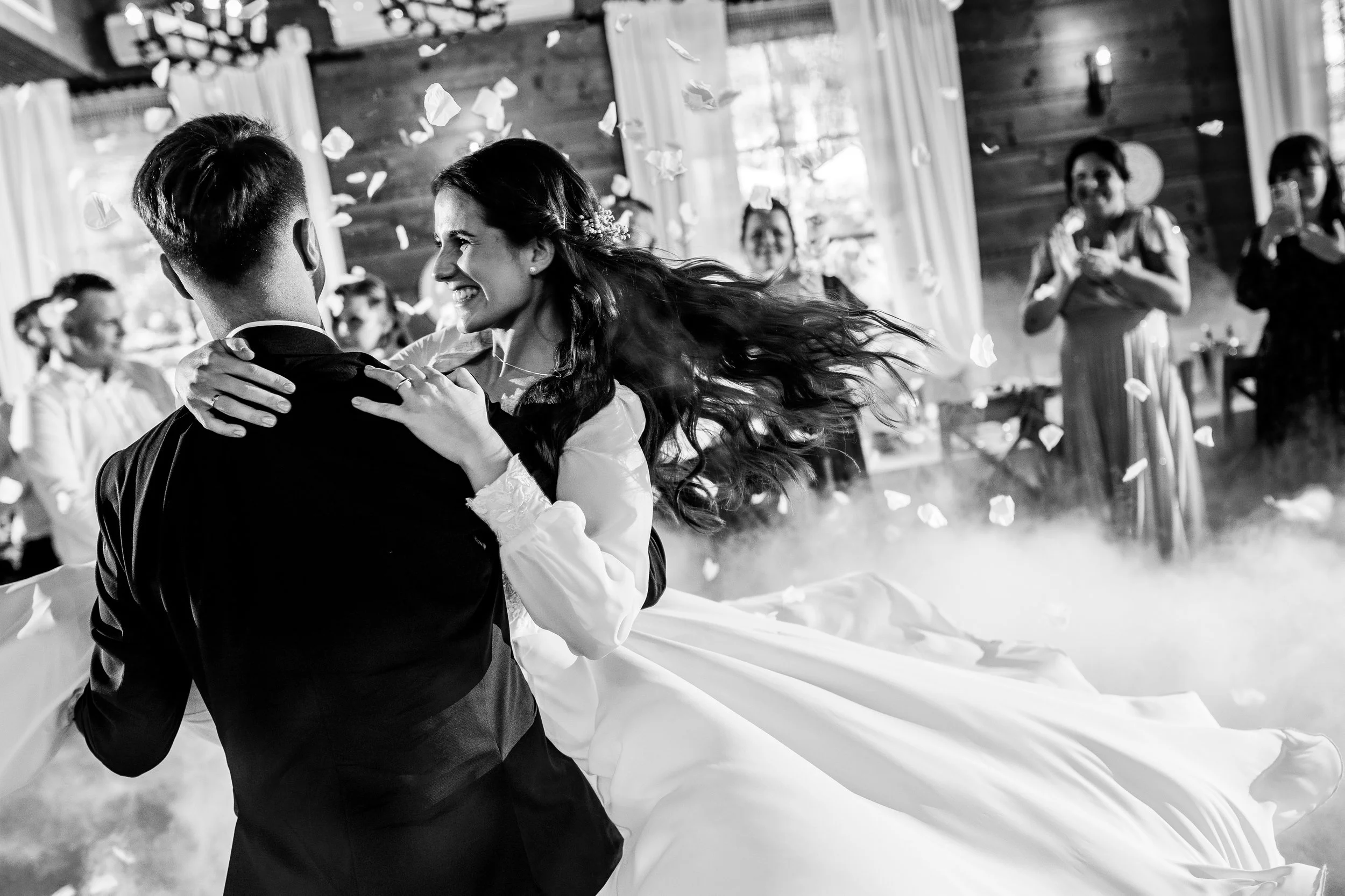 A bride and groom are dancing at their wedding reception, surrounded by guests. The woman is smiling with her hair flowing, and the man is holding her. Confetti is falling, and there is a mist or smoke on the floor in a decorated indoor venue.