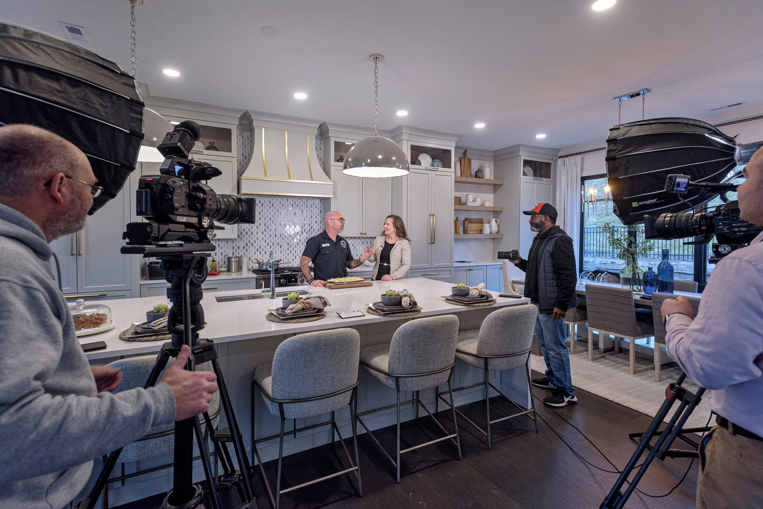 A behind-the-scenes view of a kitchen filming setup, featuring a well-lit modern kitchen with people, cameras, and lighting equipment in action.