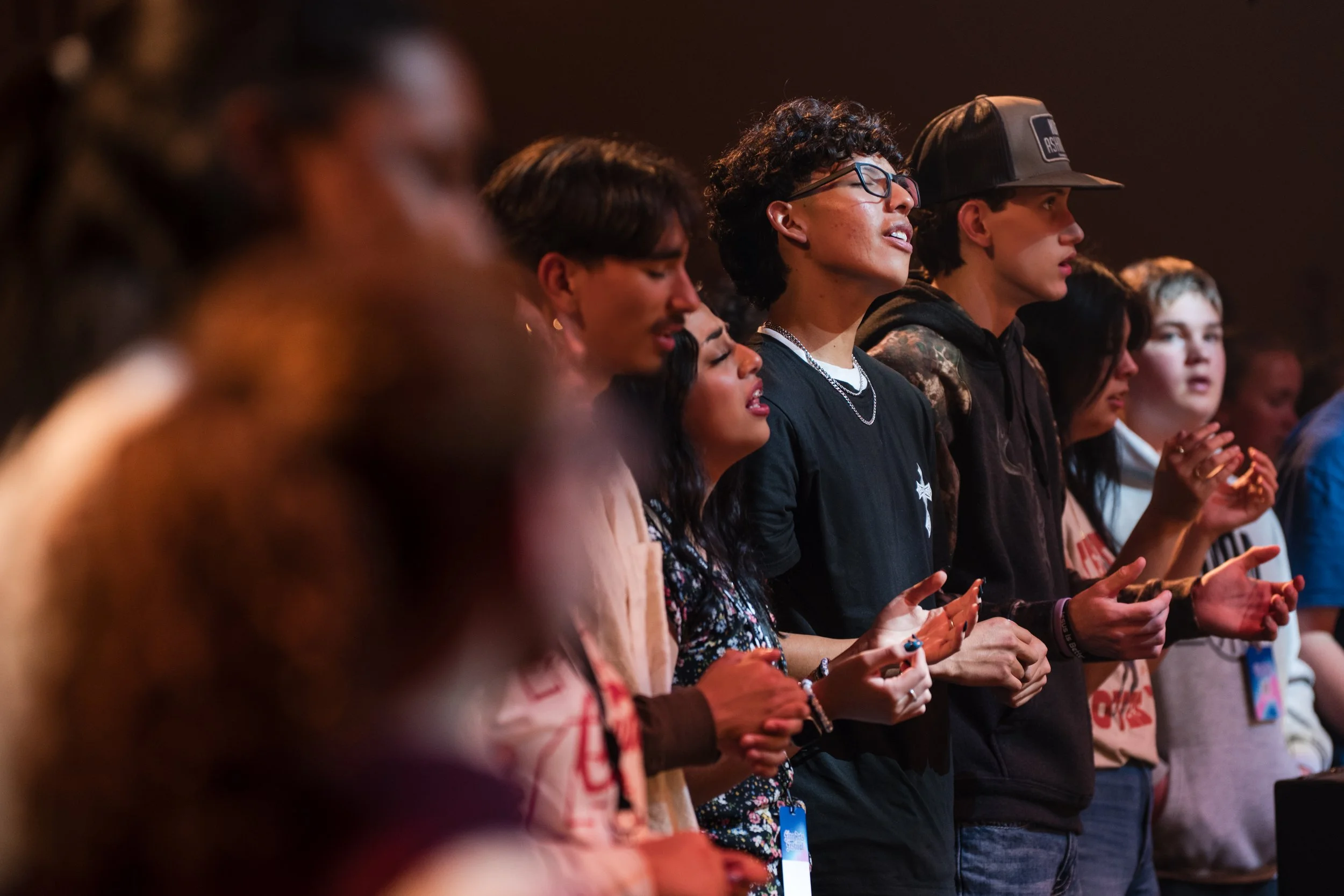 Group of young people praying or singing with eyes closed, standing in a line at a concert or event.