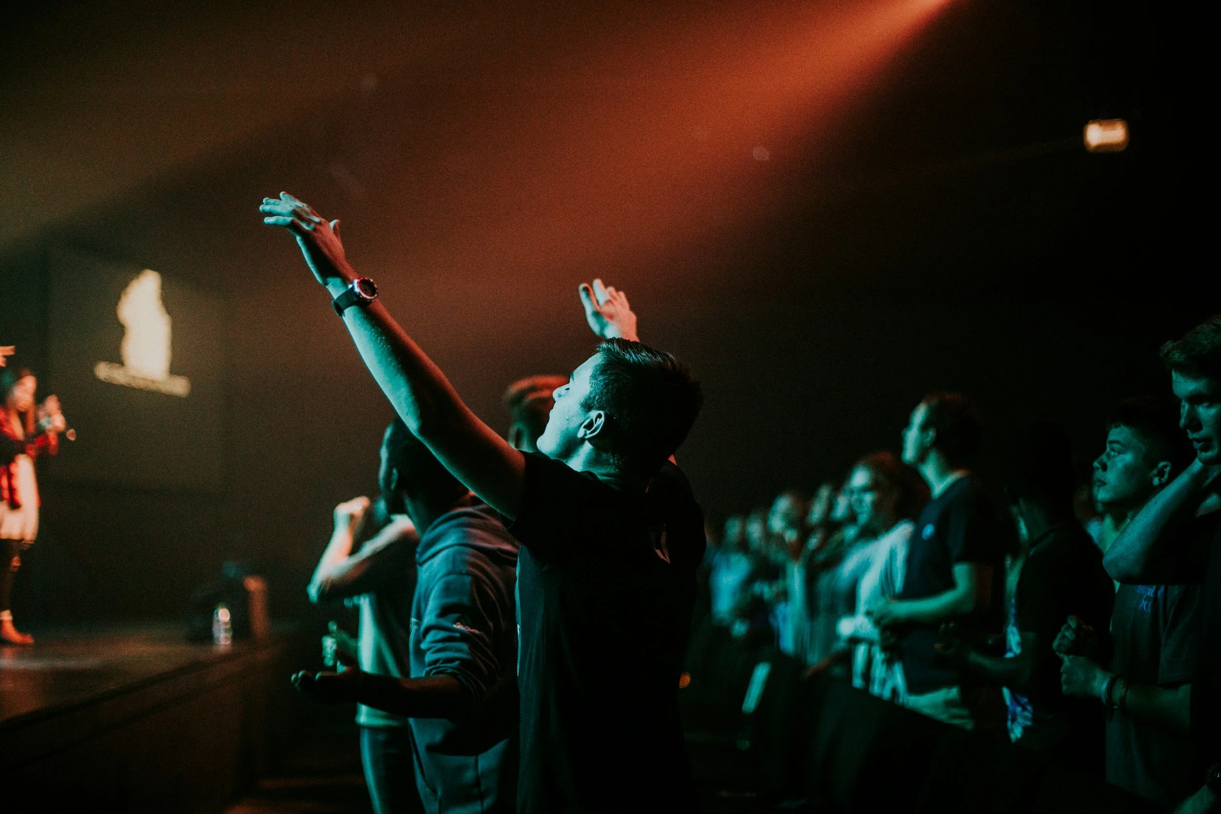 People attending a worship service or concert in a dark auditorium, some with arms raised, during a performance on stage.