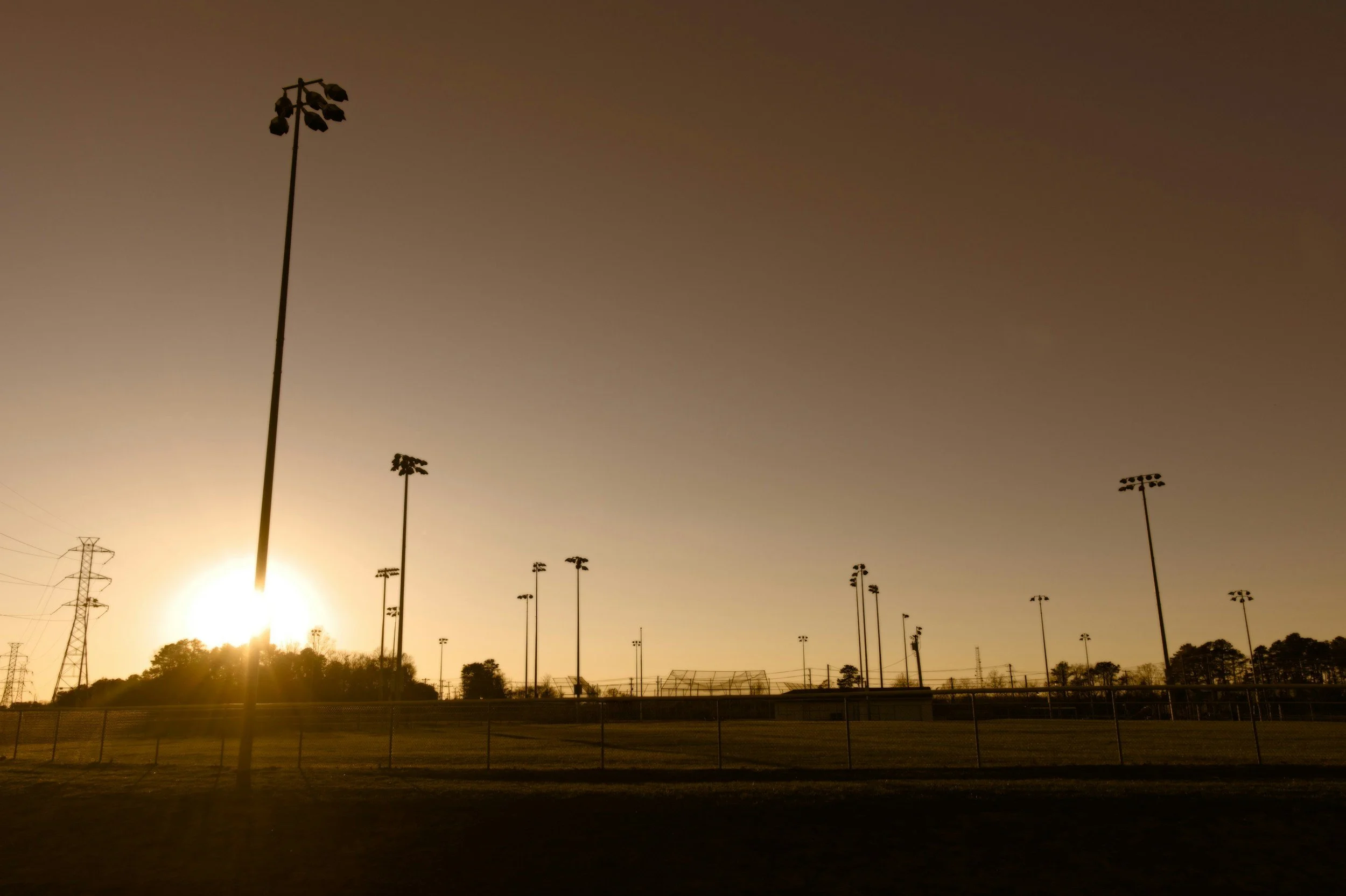 Empty softball field at sunset with tall light poles and a chain-link fence, silhouettes of trees in the background.