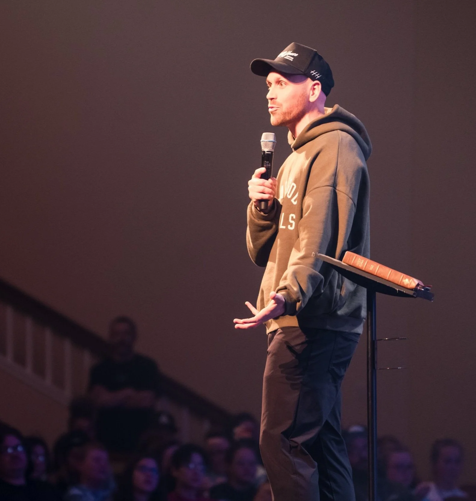 A man in a brown hoodie and black cap speaking into a microphone on stage during a presentation, with an audience watching.
