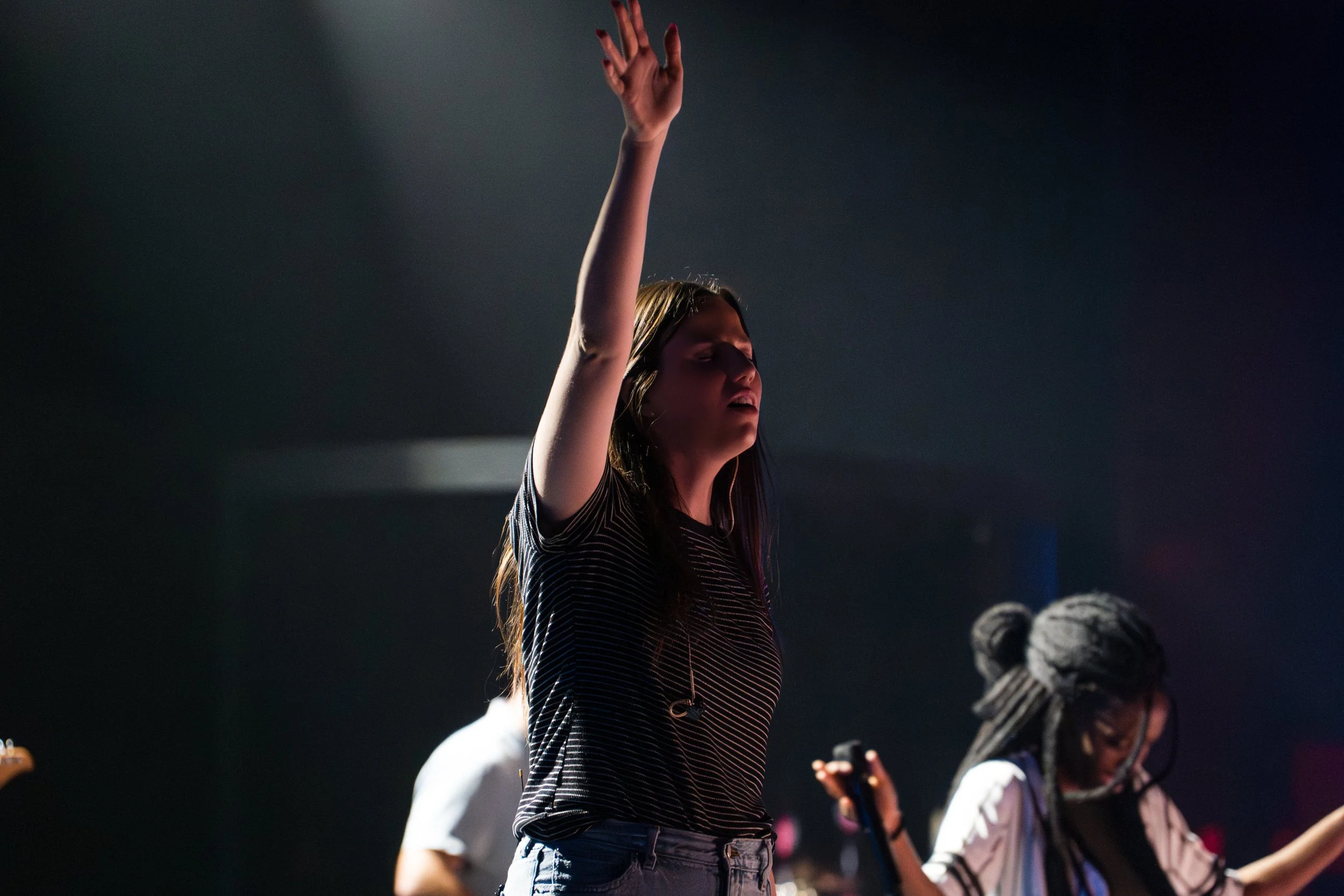 A woman with long brown hair raising her right arm in a dark room, likely at a concert or worship event, with other people visible in the background.