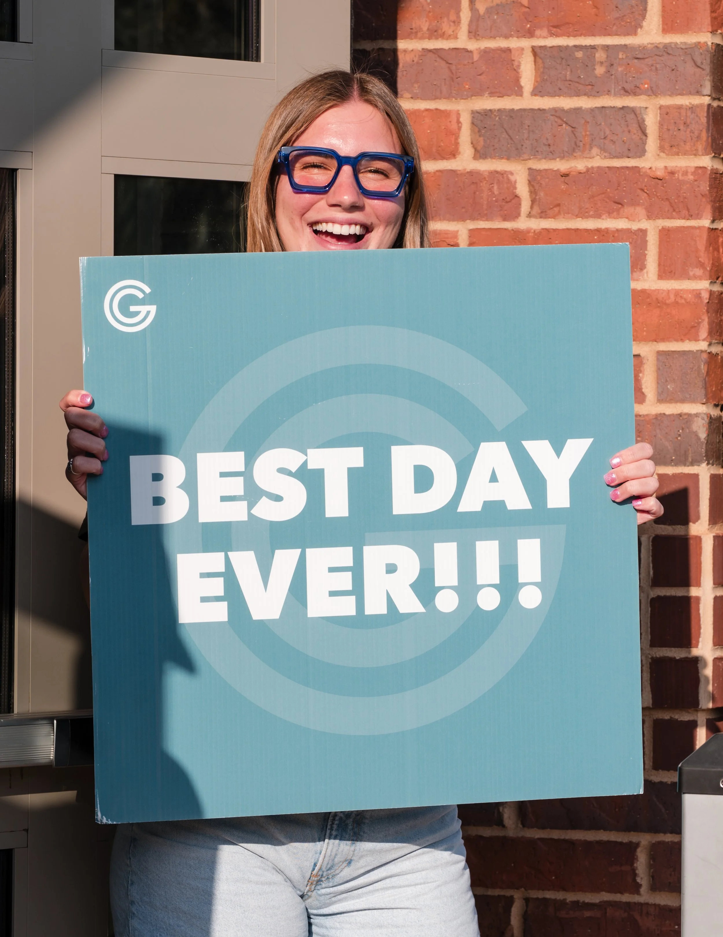A woman with blonde hair, wearing blue glasses, holding a sign that reads "Best Day Ever!!" and smiling outdoors against a brick wall.