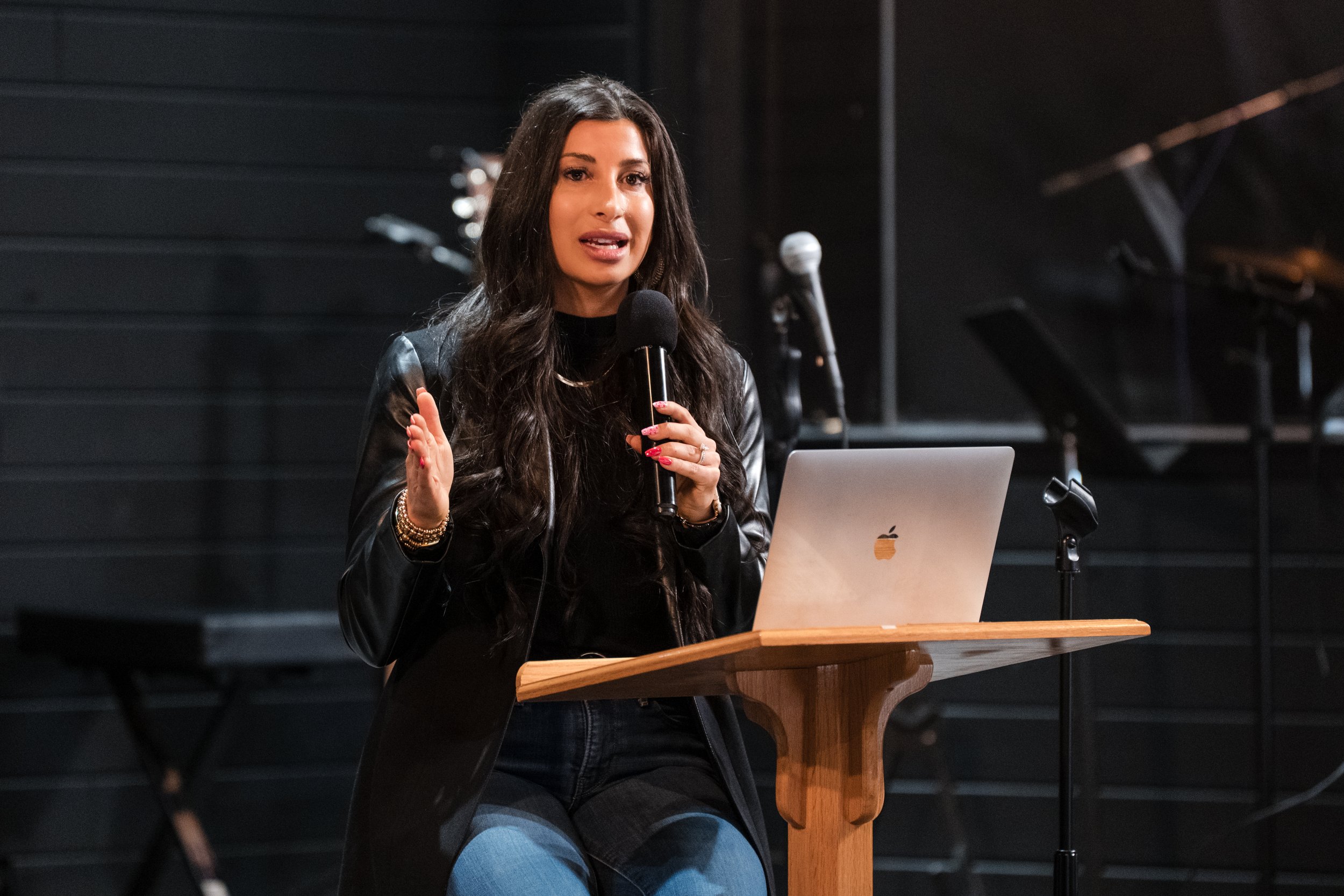 A woman with long dark hair wearing a black leather jacket and jeans, speaking into a microphone while seated at a wooden table with a silver laptop, in front of a black background with music stands and microphones.
