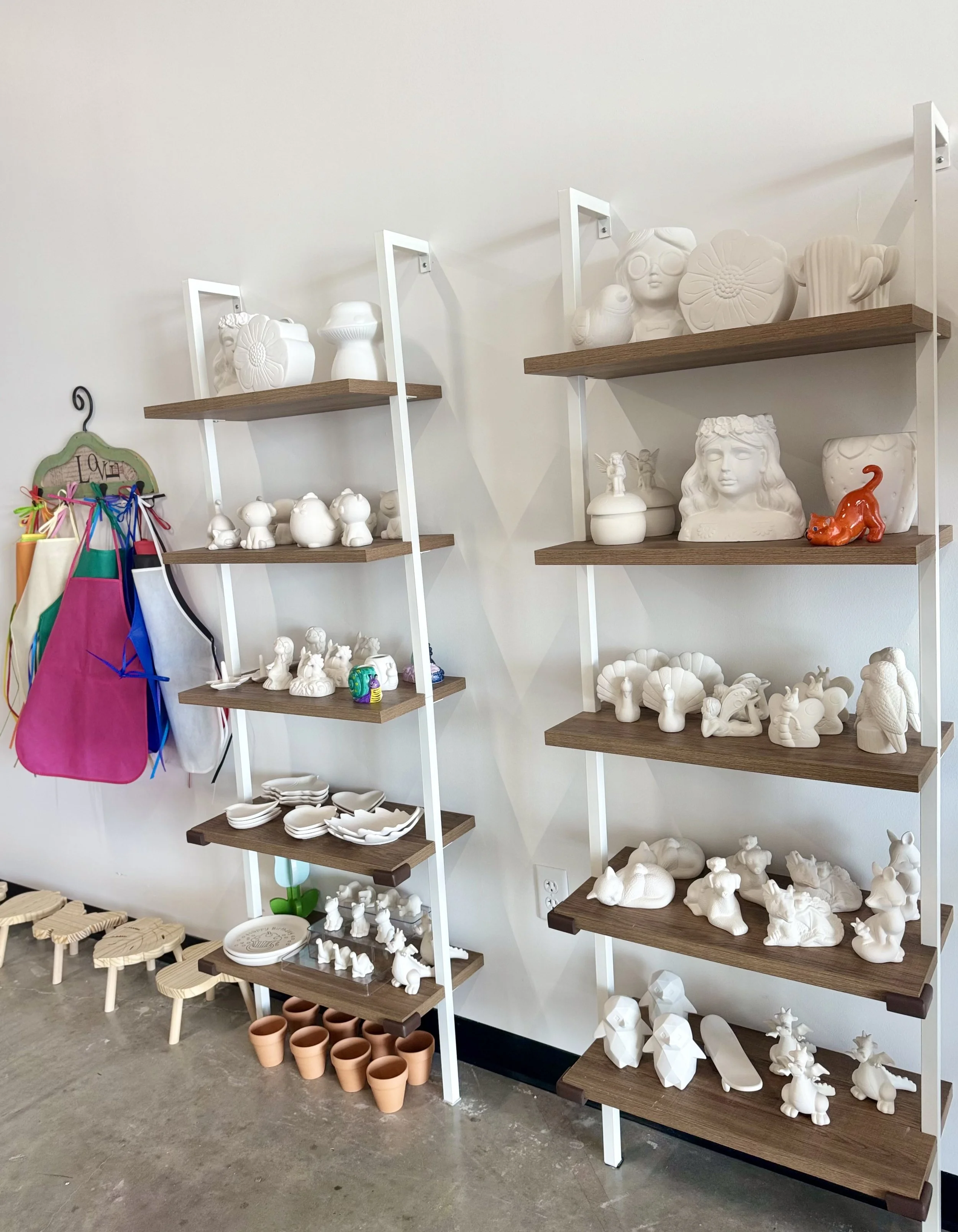 Display shelves filled with white ceramic figurines, vases, and pottery in a shop, with colorful aprons hanging on the left side.