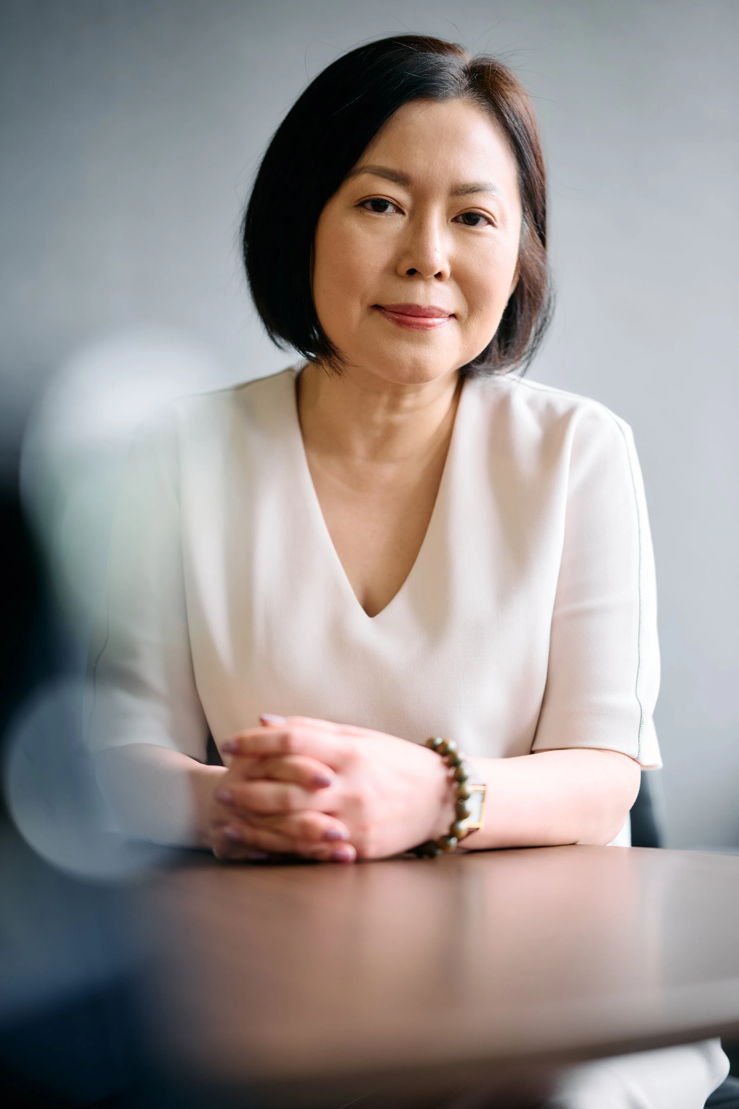 A woman with short dark hair, wearing a light-colored blouse, sitting at a table with her hands folded, looking at the camera, smiling gently.