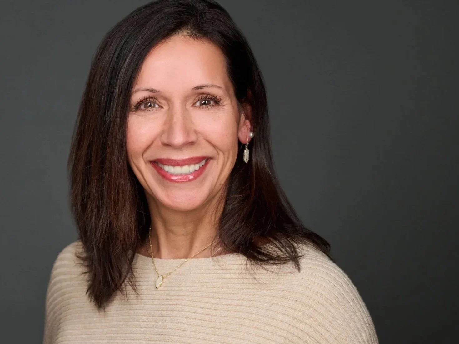 A woman with shoulder-length dark brown hair smiling at the camera, wearing a beige sweater, pearl earrings, and a gold necklace with a heart-shaped pendant against a dark gray background.