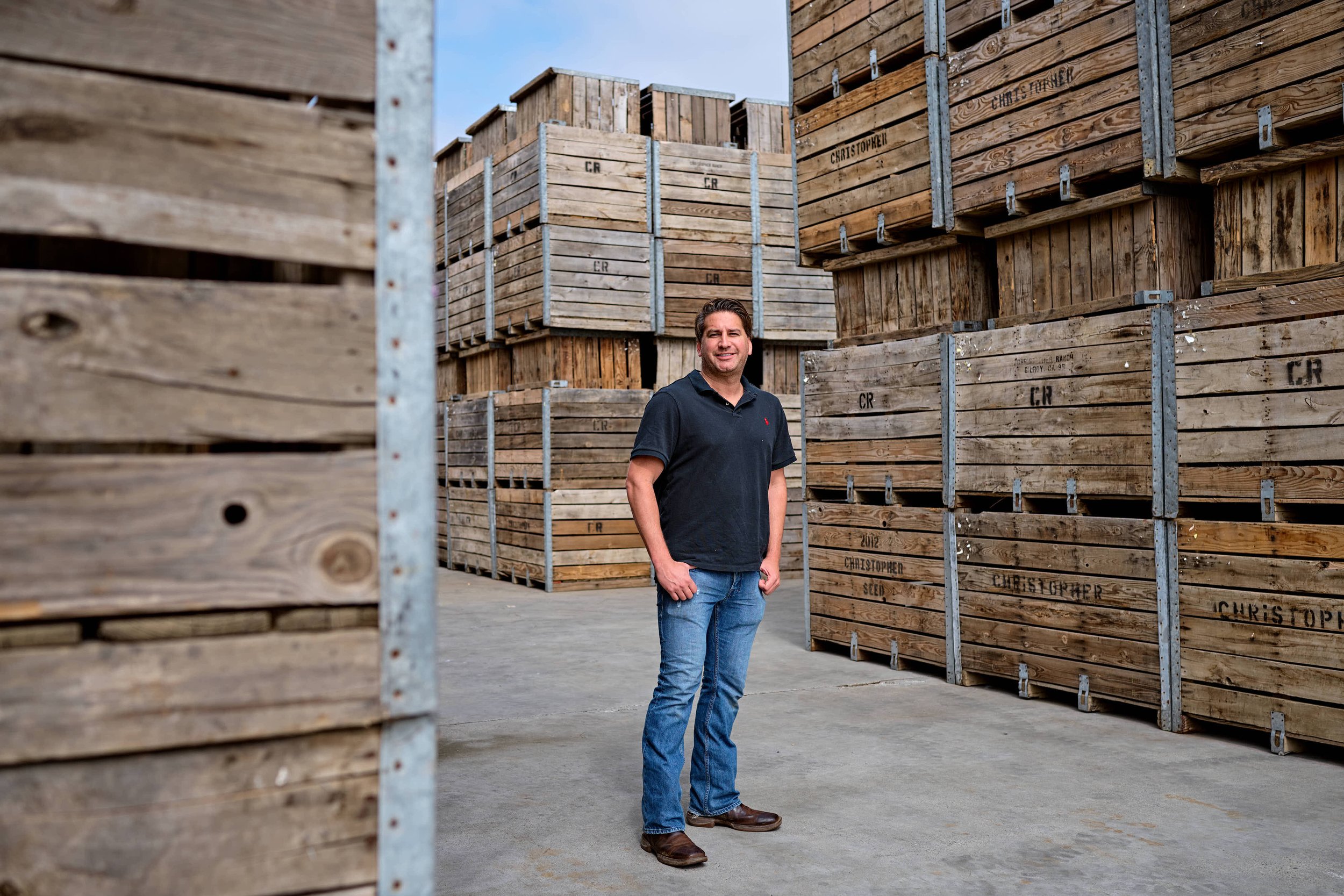 A man standing in an outdoor warehouse or lumber yard surrounded by stacked large wooden crates.