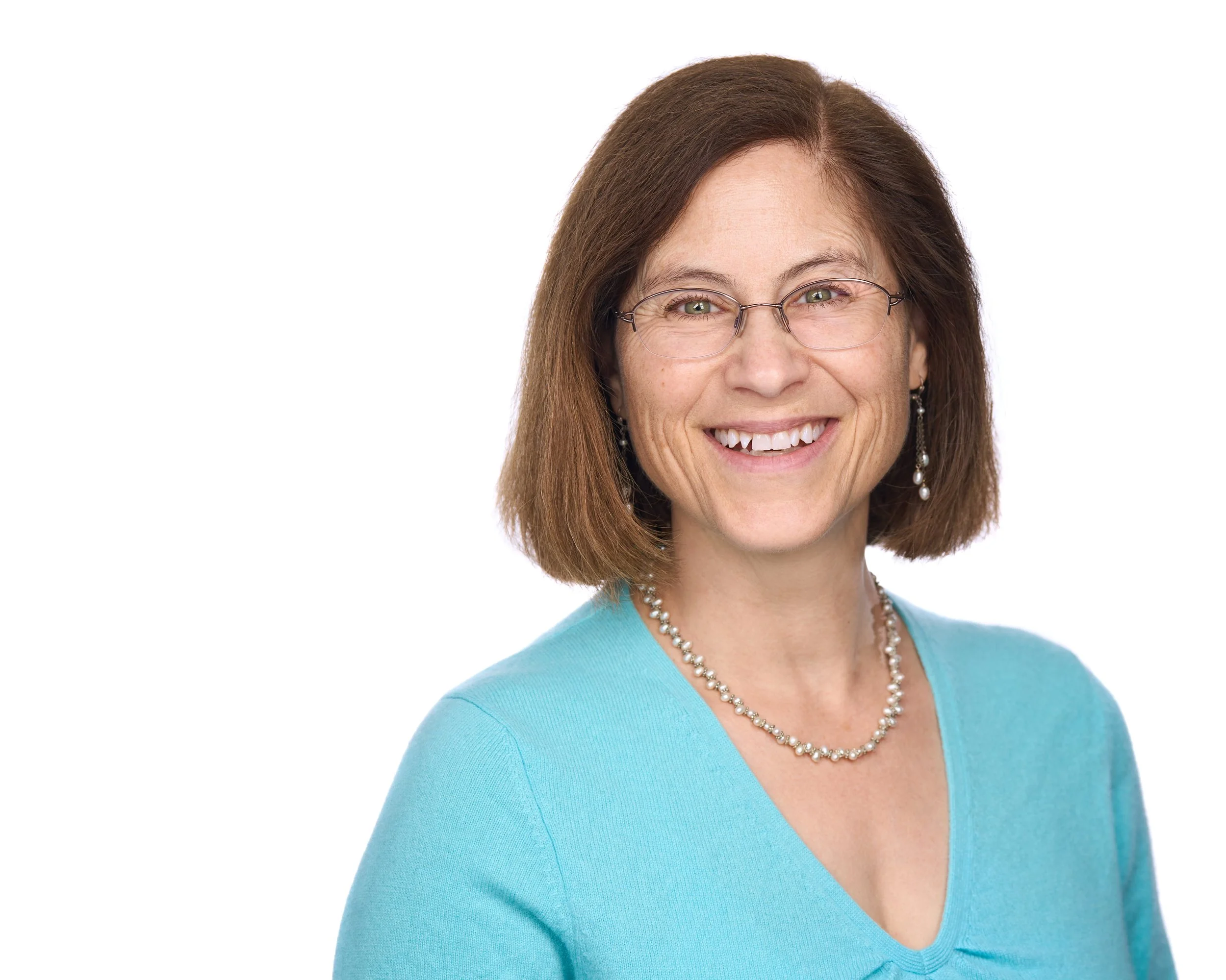 A smiling woman with brown hair, glasses, and earrings, wearing a turquoise top and a pearl necklace, against a plain white background.