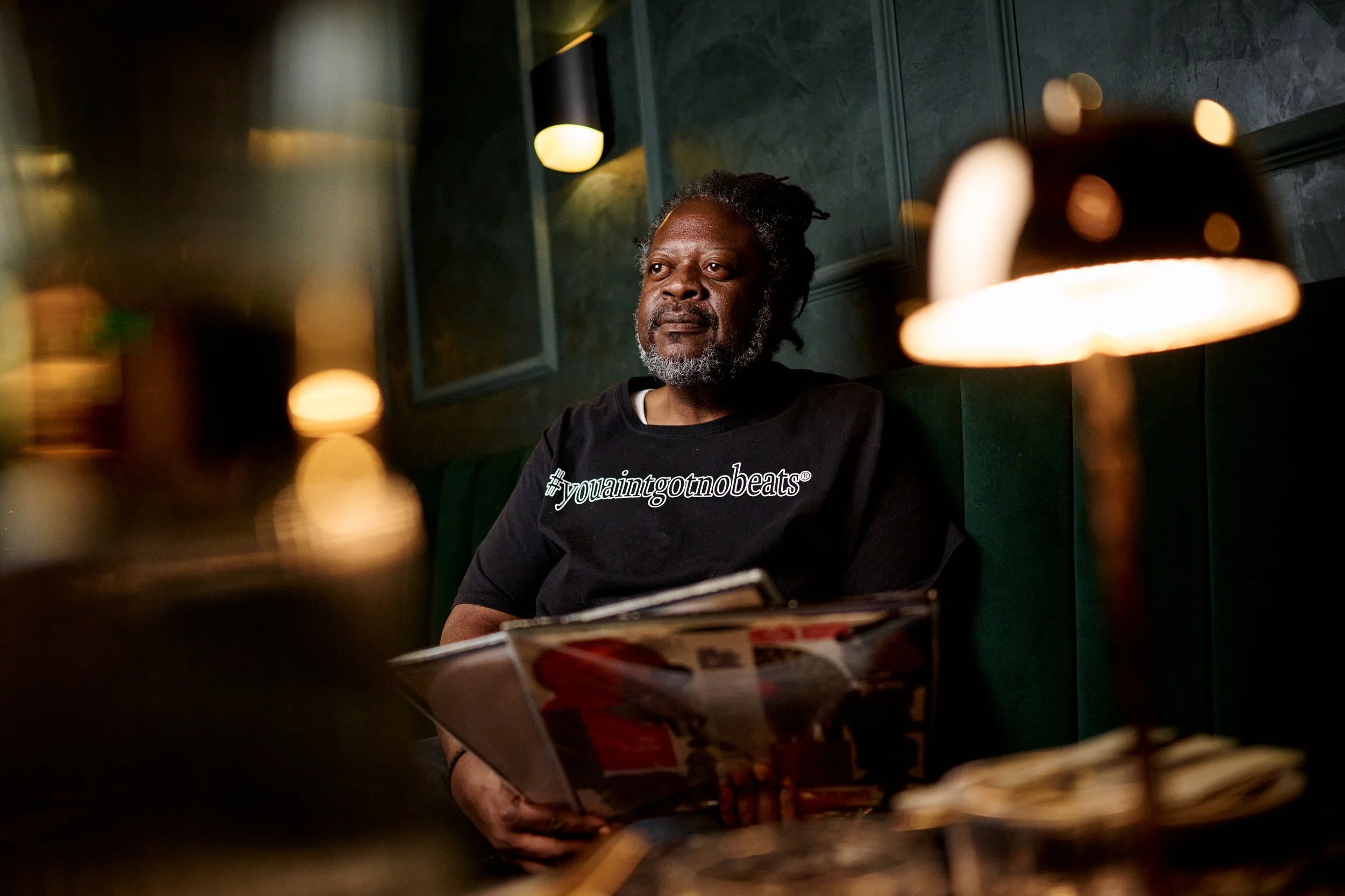 A man with gray hair and a beard sitting in a dimly lit room, wearing a black t-shirt with '#youaintgonnobeats' written on it, holding a magazine.